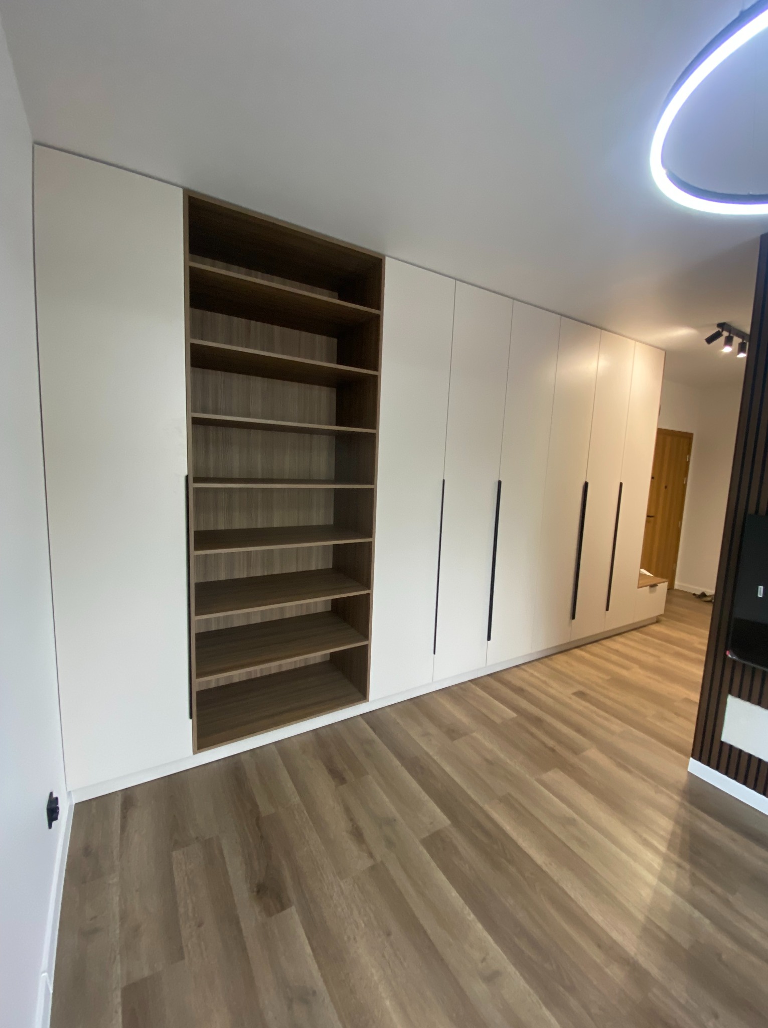 Biała zabudowa meblowa na wymiar z drewnianymi półkami i długimi, czarnymi uchwytami, widok z perspektywy podłogi,  oświetlenie sufitowe LED.
