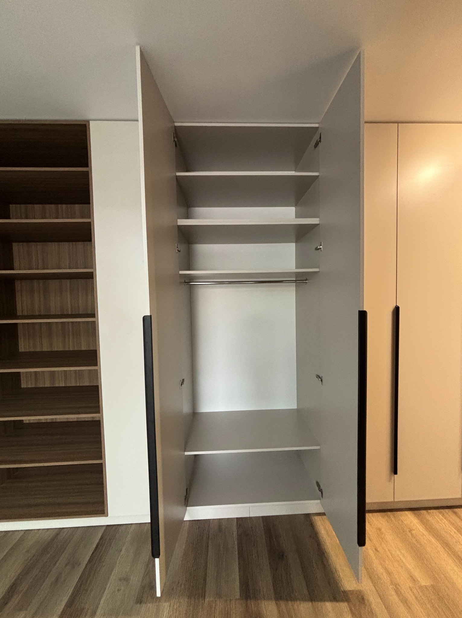 Wnętrze szafy na wymiar z otwartymi drzwiami, widoczne półki i drążek na ubrania. Obok regał z półkami. Minimalistyczny design, jasne kolory.