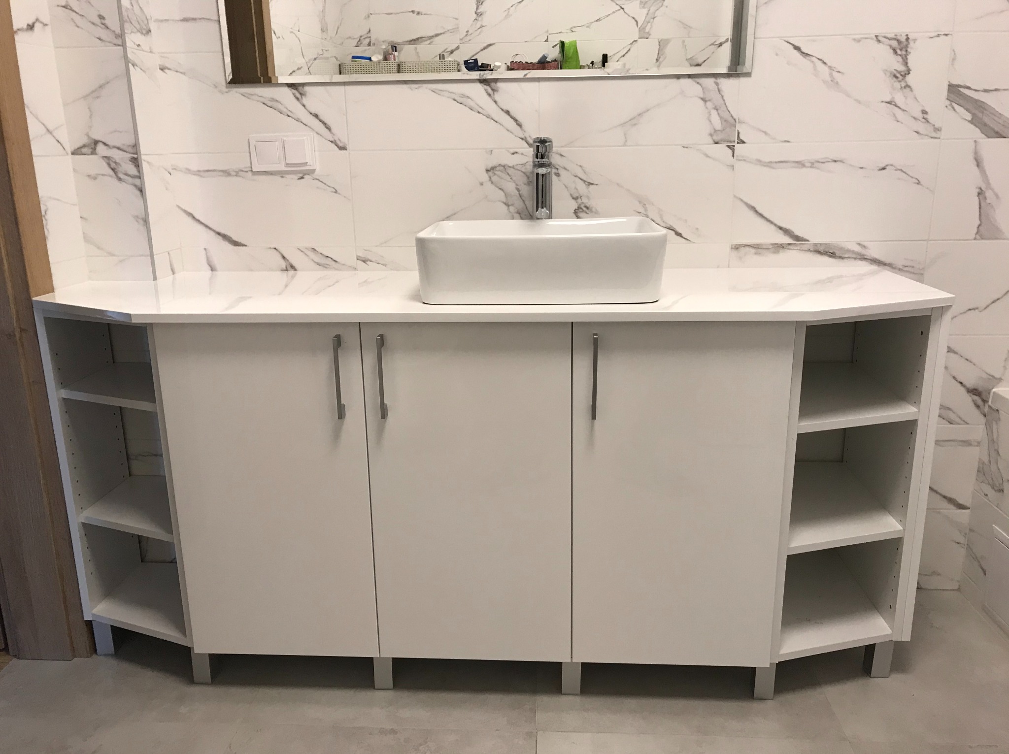 Biała szafka łazienkowa pod umywalkę z blatem imitującym marmur, kwadratowa umywalka nablatowa, srebrna bateria, w tle marmurowe płytki.