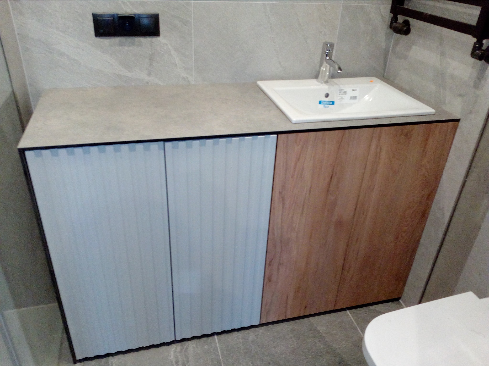 Szafka łazienkowa z umywalką: fronty w odcieniach błękitu i drewna, szary blat, płytki imitujące marmur w tle, nowoczesna armatura.