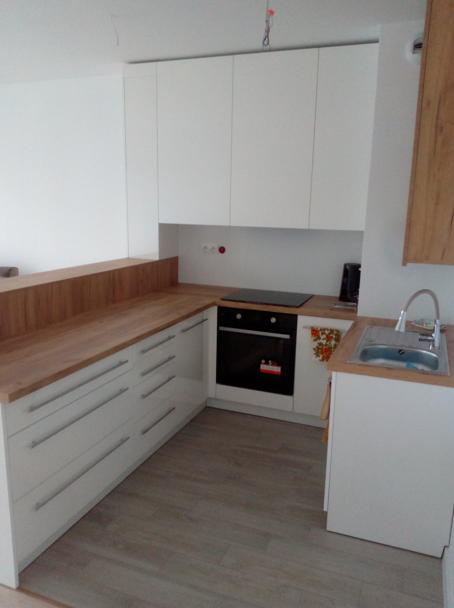 Nowoczesna, biała kuchnia z drewnianym blatem, widok na zlewozmywak, płytę grzewczą i piekarnik. Minimalistyczny design z długimi, poziomymi uchwytami.