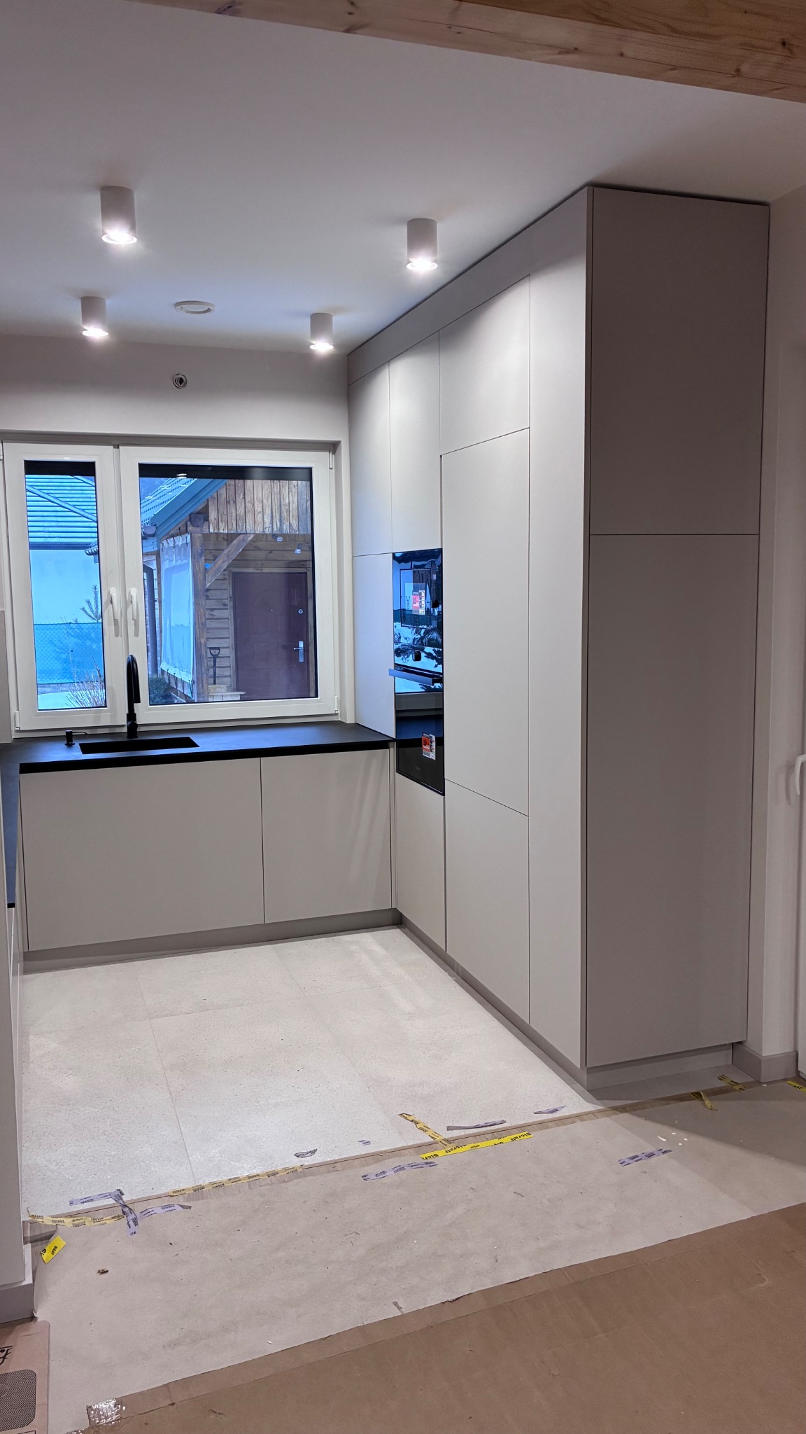 Jasna, minimalistyczna kuchnia z zabudową pod sufit, czarnym blatem i widokiem na drewniany budynek przez okno. Podłoga wyłożona jasnymi płytkami, widoczne ślady po remoncie.