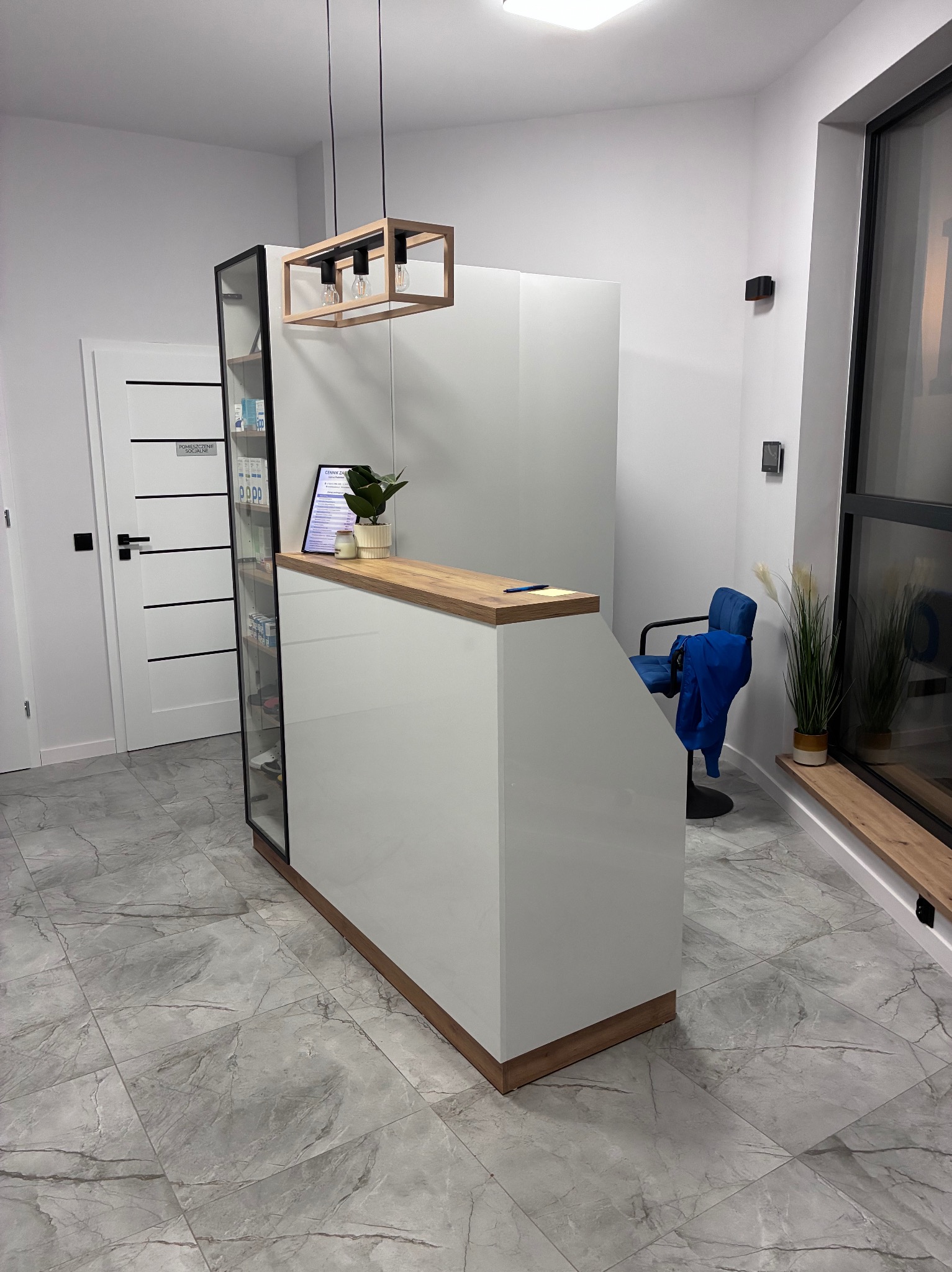 Biała lada recepcyjna z drewnianym blatem i oświetleniem w nowoczesnym wnętrzu. Na blacie roślina w doniczce. W tle krzesło i okno.