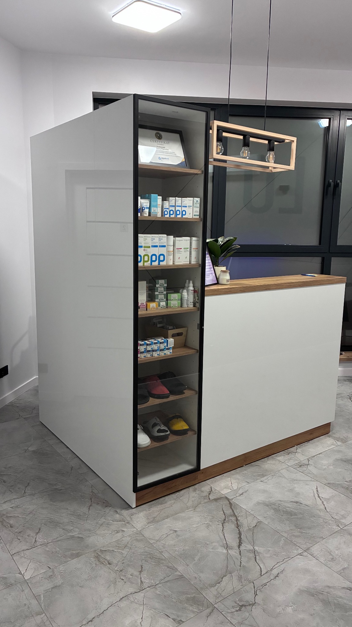 Biała lada recepcyjna z przeszkloną witryną, w której znajdują się produkty i certyfikat. Uwagę przykuwa nowoczesna lampa wisząca i marmurowa podłoga.