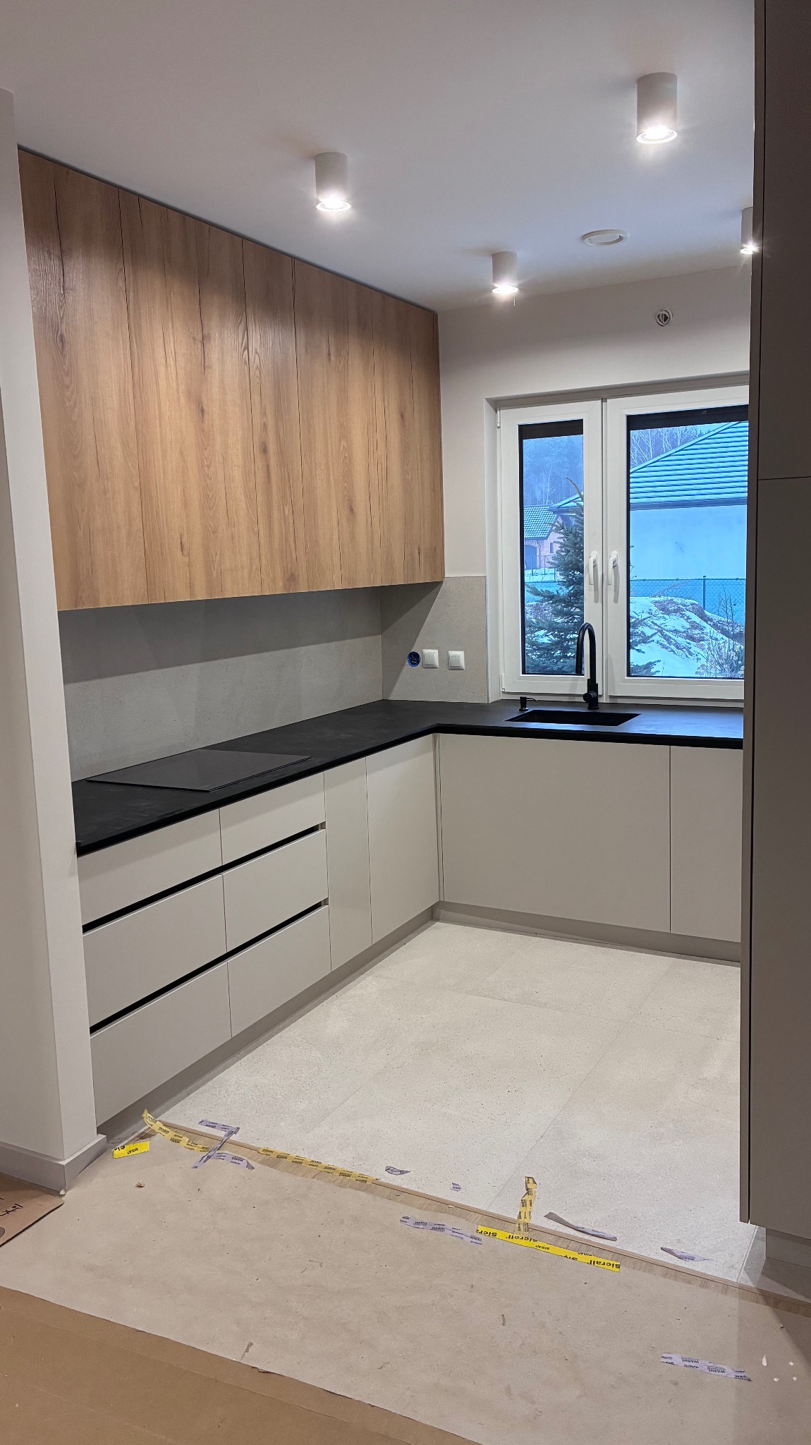 Jasna kuchnia z drewnianymi szafkami wiszącymi i czarnym blatem, widok na zimowy krajobraz przez okno. Na podłodze widoczne taśmy zabezpieczające.