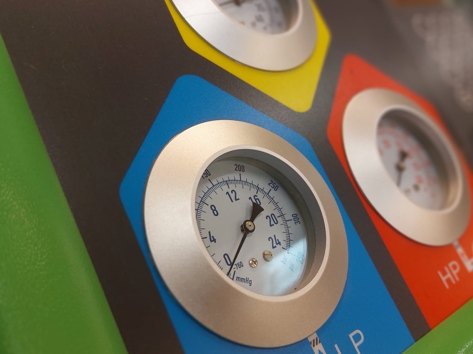 Manometry w kolorowych obudowach (żółta, niebieska, czerwona, zielona) na panelu urządzenia do serwisowania klimatyzacji samochodowej. Widok z bliska, z boku.