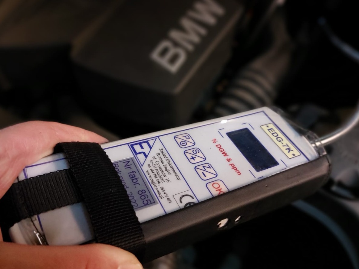 Tester szczelności klimatyzacji samochodowej EDG-7K* w dłoni, na tle silnika BMW. Pomiar % DGW i ppm, rok produkcji 2022.