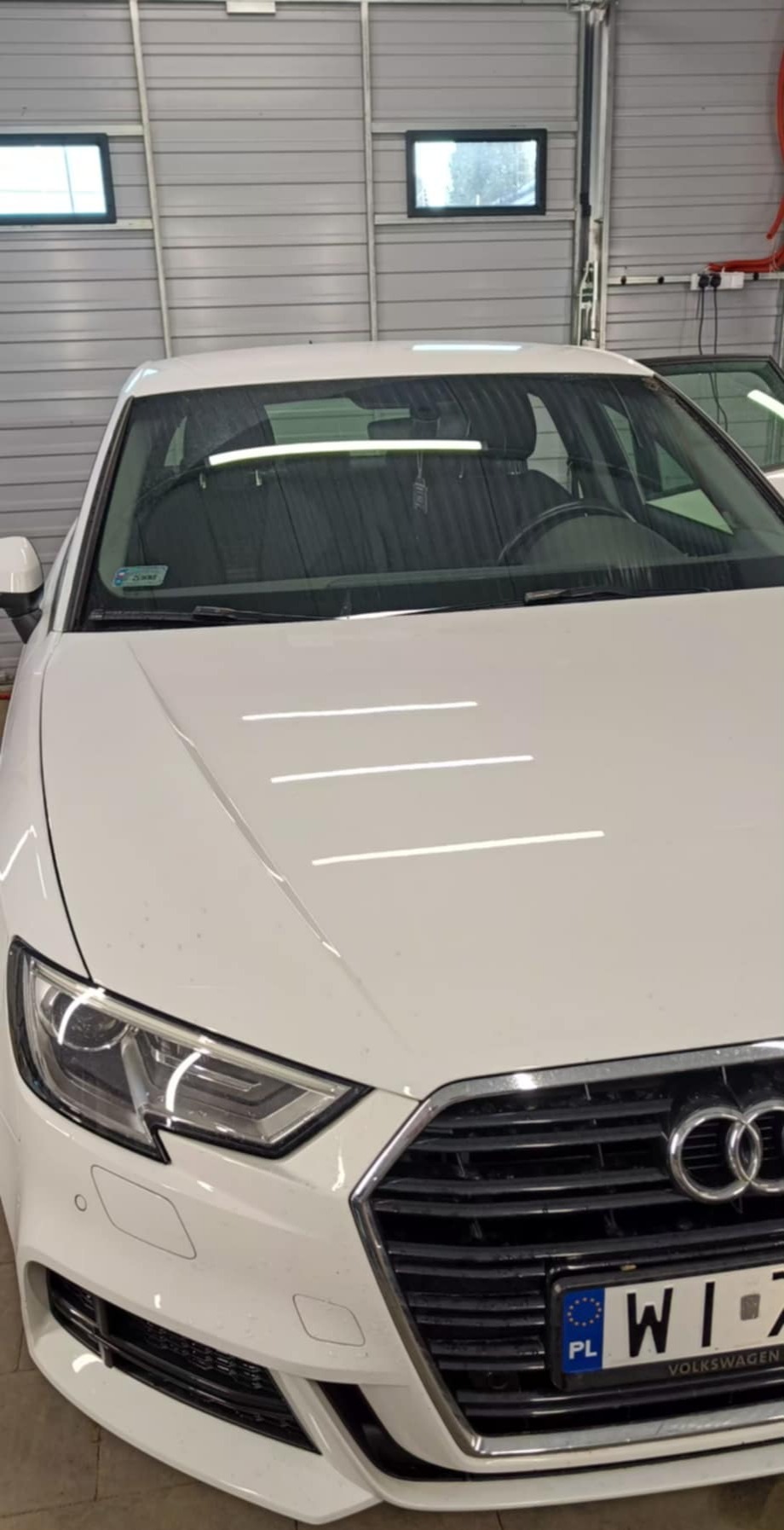 Białe Audi A3 w warsztacie z widoczną tablicą rejestracyjną, ukazujące detale karoserii i reflektorów, sugerujące mycie lub detailing.