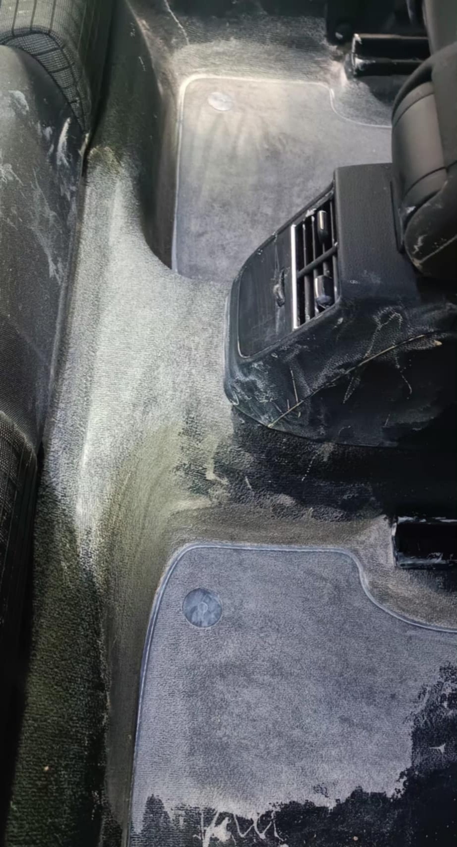 Wnętrze samochodu z wypraną, ale jeszcze mokrą tapicerką podłogową, widoczne ślady detergentu i pozostałości po czyszczeniu na dywanikach i plastikowych elementach.