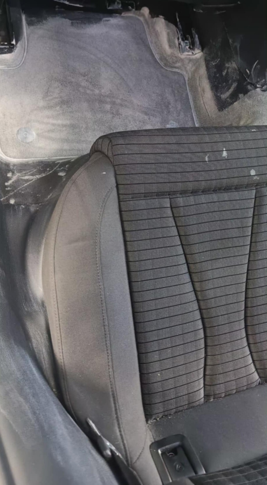 Wnętrze auta z widocznym, częściowo wyczyszczonym fotelem z tapicerką w kratę i fragmentem podłogi pokrytej środkiem czyszczącym. Widoczny etap czyszczenia.