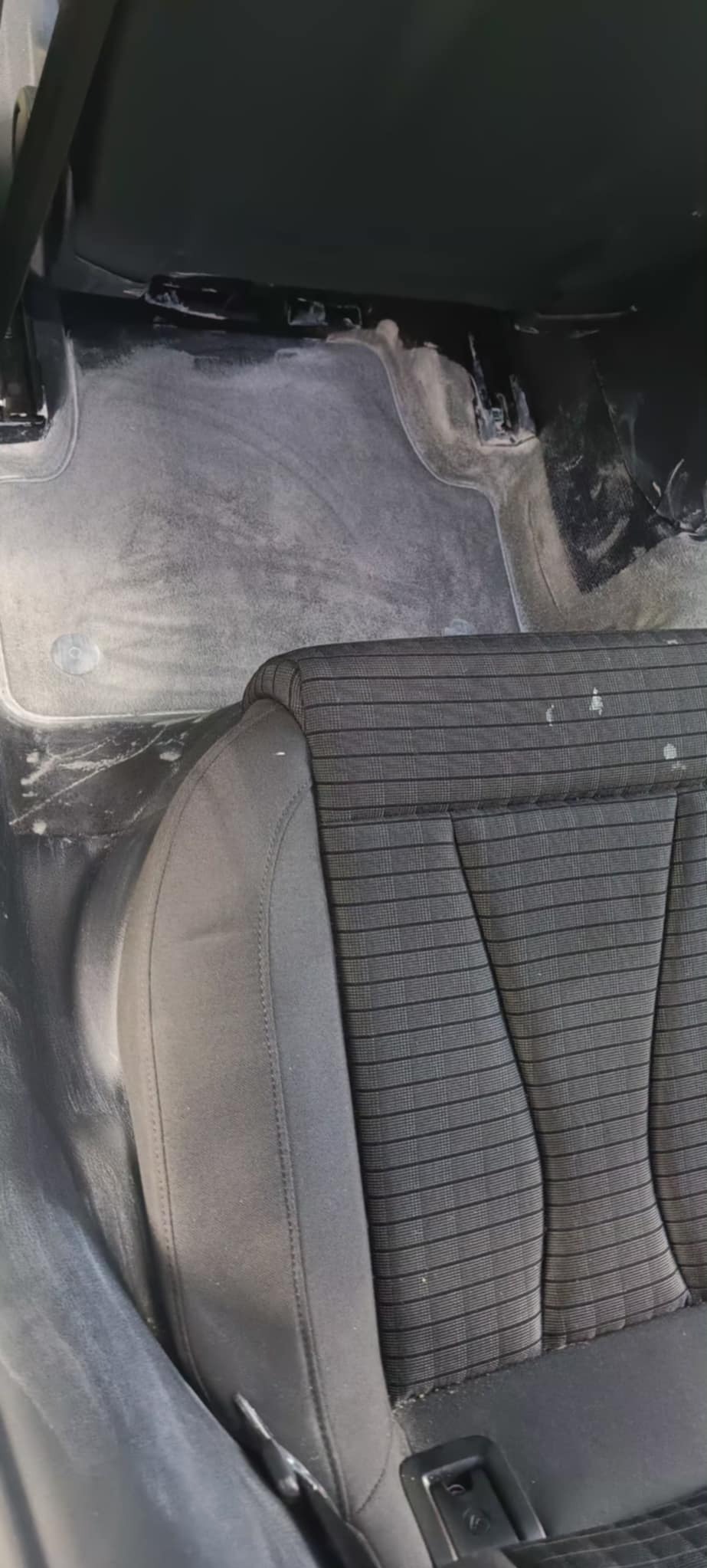 Wnętrze samochodu w trakcie czyszczenia: fotel z tapicerką w kratę i podłoga pokryta pianą czyszczącą. Widoczne zabrudzenia na tapicerce.