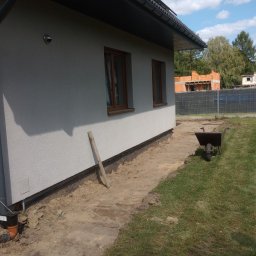 Kopman Robert Palewicz - Przygotowanie terenu wokół domu pod drenaż, widoczne odkryte fundamenty z siatką, rura odpływowa, taczka i drewniana listwa oparta o ścianę.