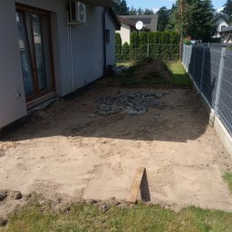 Kopman Robert Palewicz - Przygotowany teren pod budowę tarasu: wyrównana powierzchnia z piasku, pryzma kamieni, deska oparta o brzeg, widok z góry na tle domu i ogrodzenia.