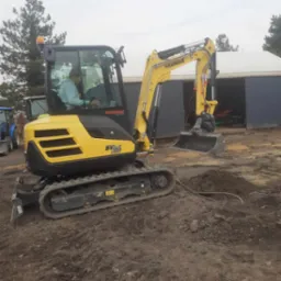 Żółta minikoparka Yanmar SV26 w trakcie pracy, widoczne wykopane zagłębienie w ziemi i wystające rury, operator w kabinie, tło stanowią budynki gospodarcze i drzewa.