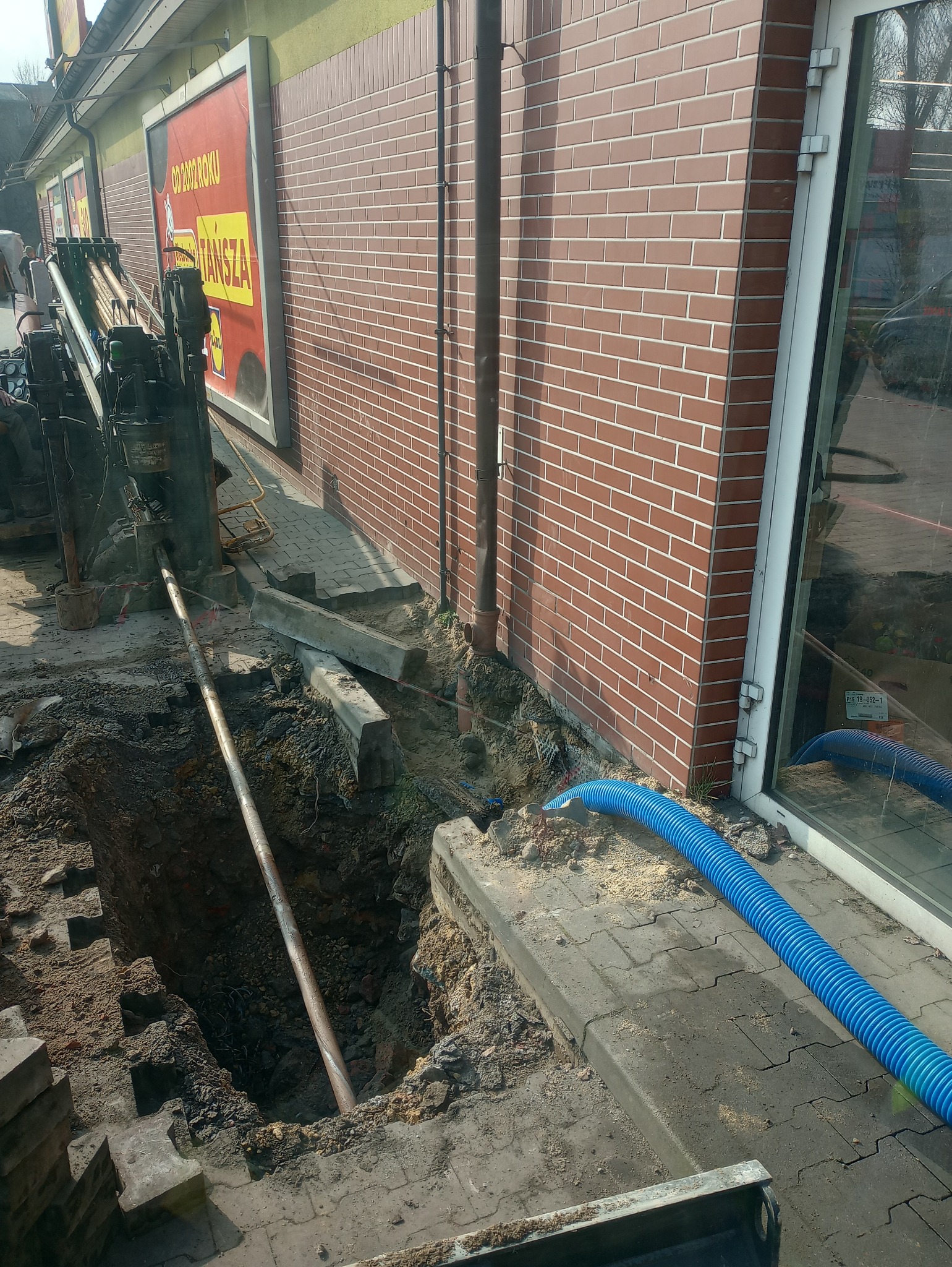 Wiertnica pozioma wykonuje odwiert pod chodnikiem w pobliżu budynku z cegły, widoczny niebieski wąż ssący i wykop.