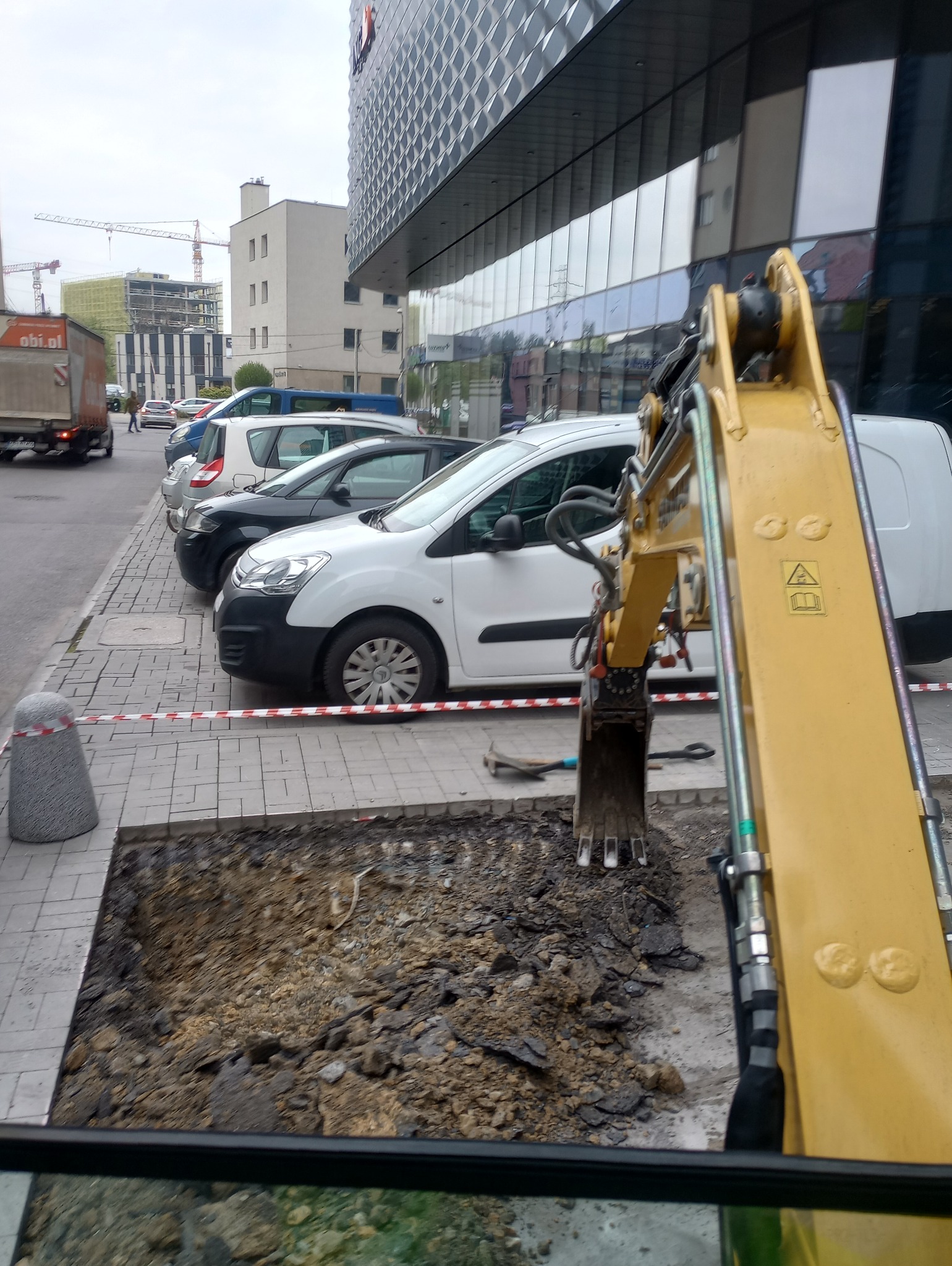 Żółta koparka z łyżką w wykopie na chodniku, w tle zaparkowane samochody i budynek biurowy, teren zabezpieczony taśmą ostrzegawczą.