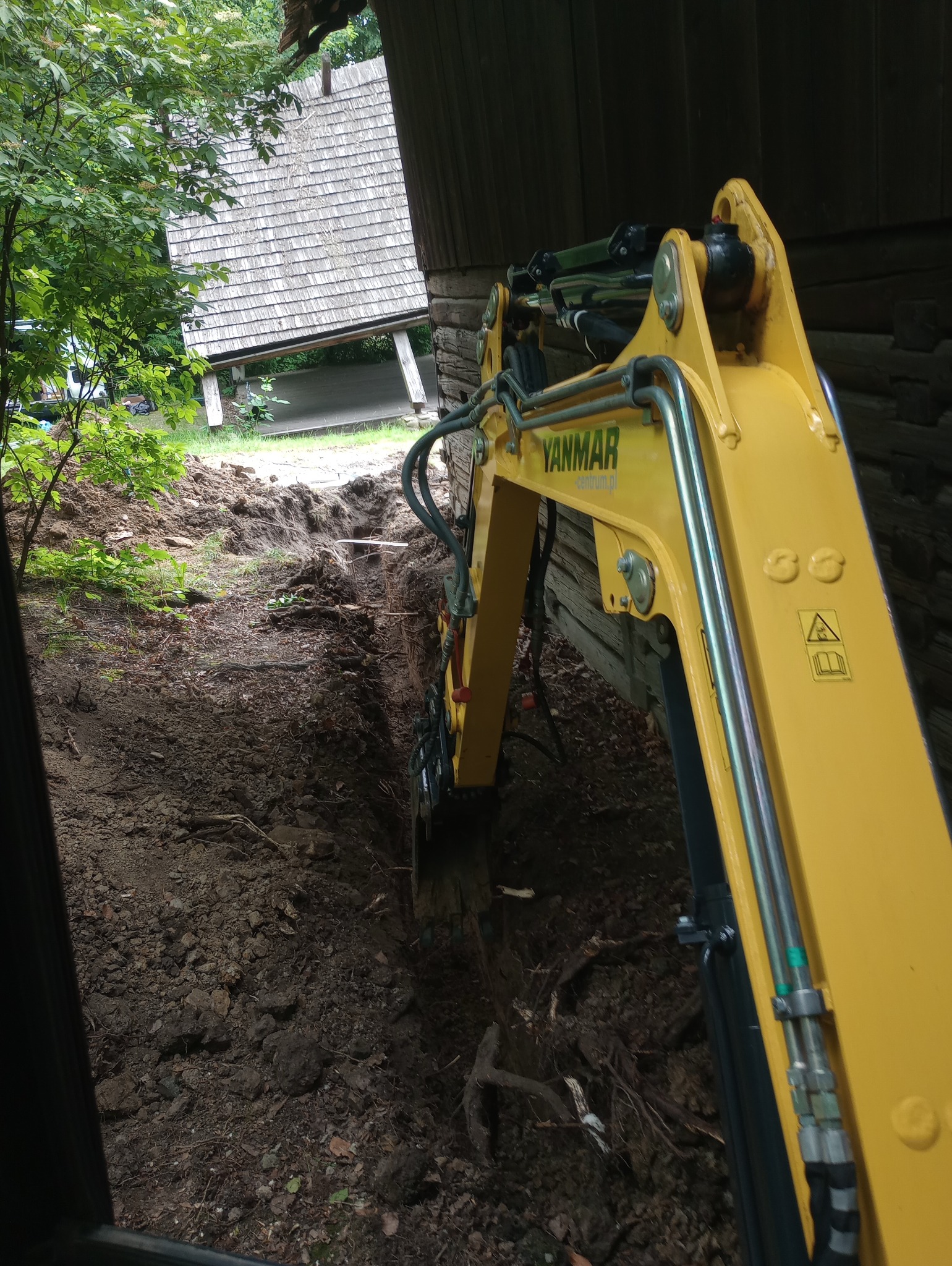 Żółta koparka Yanmar w trakcie pracy, widoczny wykop w ziemi z korzeniami, w tle drewniana zabudowa z szarym dachem.