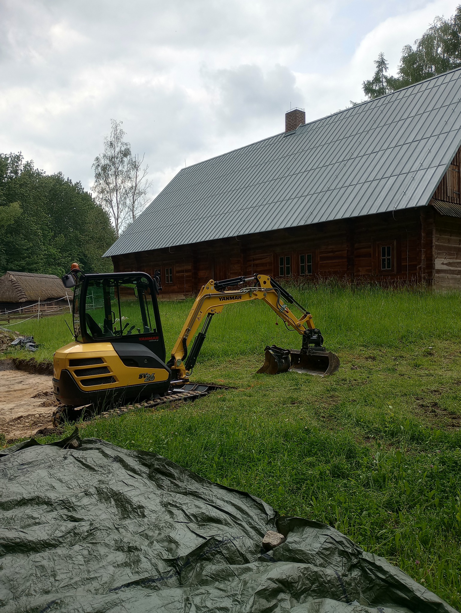 Żółta mini koparka Yanmar SV26 na trawiastym terenie, z drewnianym budynkiem w tle, obok wykopany rów, przykryty częściowo zieloną plandeką.