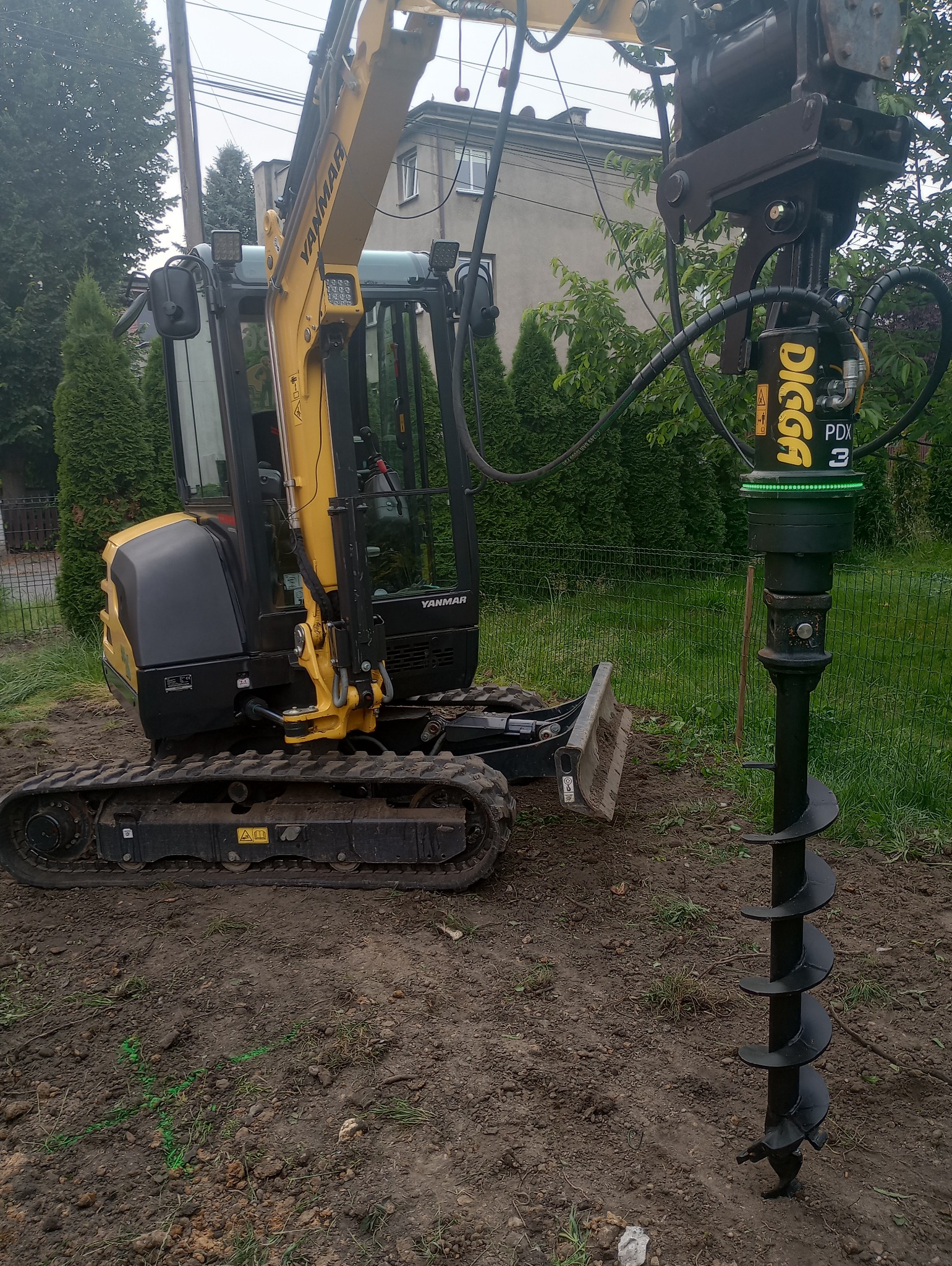 Żółto-czarna minikoparka Yanmar z wiertnicą DIGGA PDX3, wiercąca otwór w ziemi na działce z trawą i drzewami w tle.