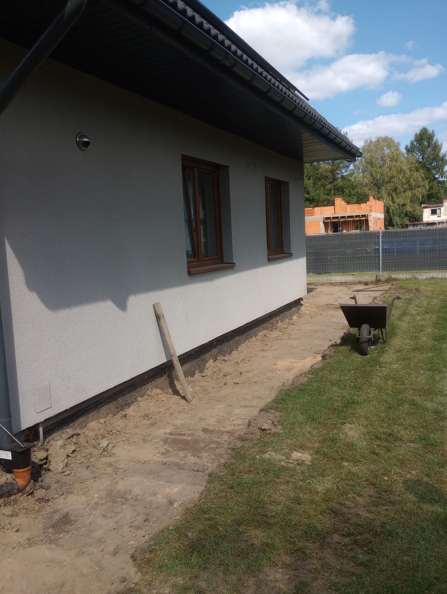 Przygotowanie terenu wokół domu pod drenaż, widoczne odkryte fundamenty z siatką, rura odpływowa, taczka i drewniana listwa oparta o ścianę.