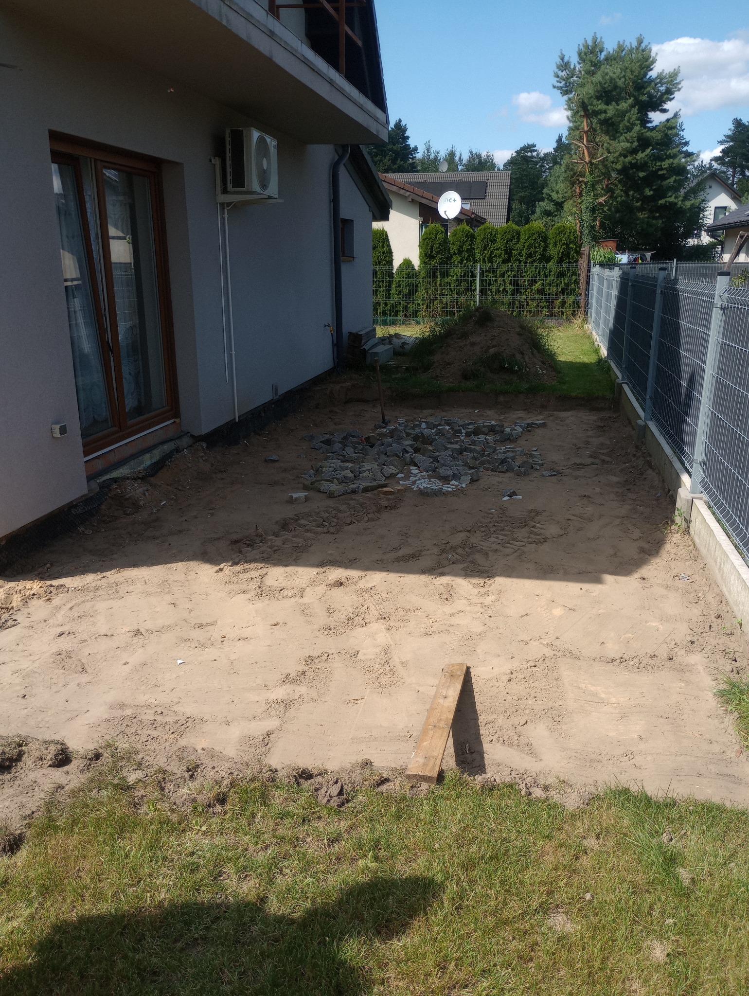 Przygotowany teren pod budowę tarasu: wyrównana powierzchnia z piasku, pryzma kamieni, deska oparta o brzeg, widok z góry na tle domu i ogrodzenia.