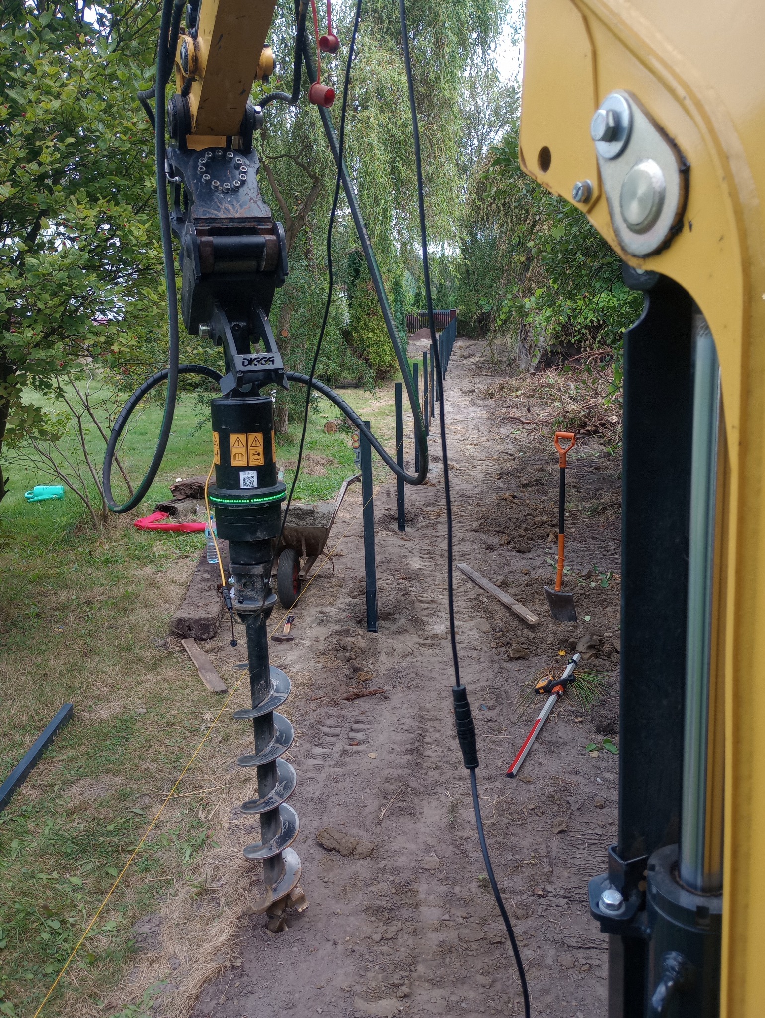 Żółta maszyna z wiertłem ślimakowym w trakcie wiercenia otworów pod słupki ogrodzenia na działce, widoczne narzędzia i częściowo wkopane słupki.