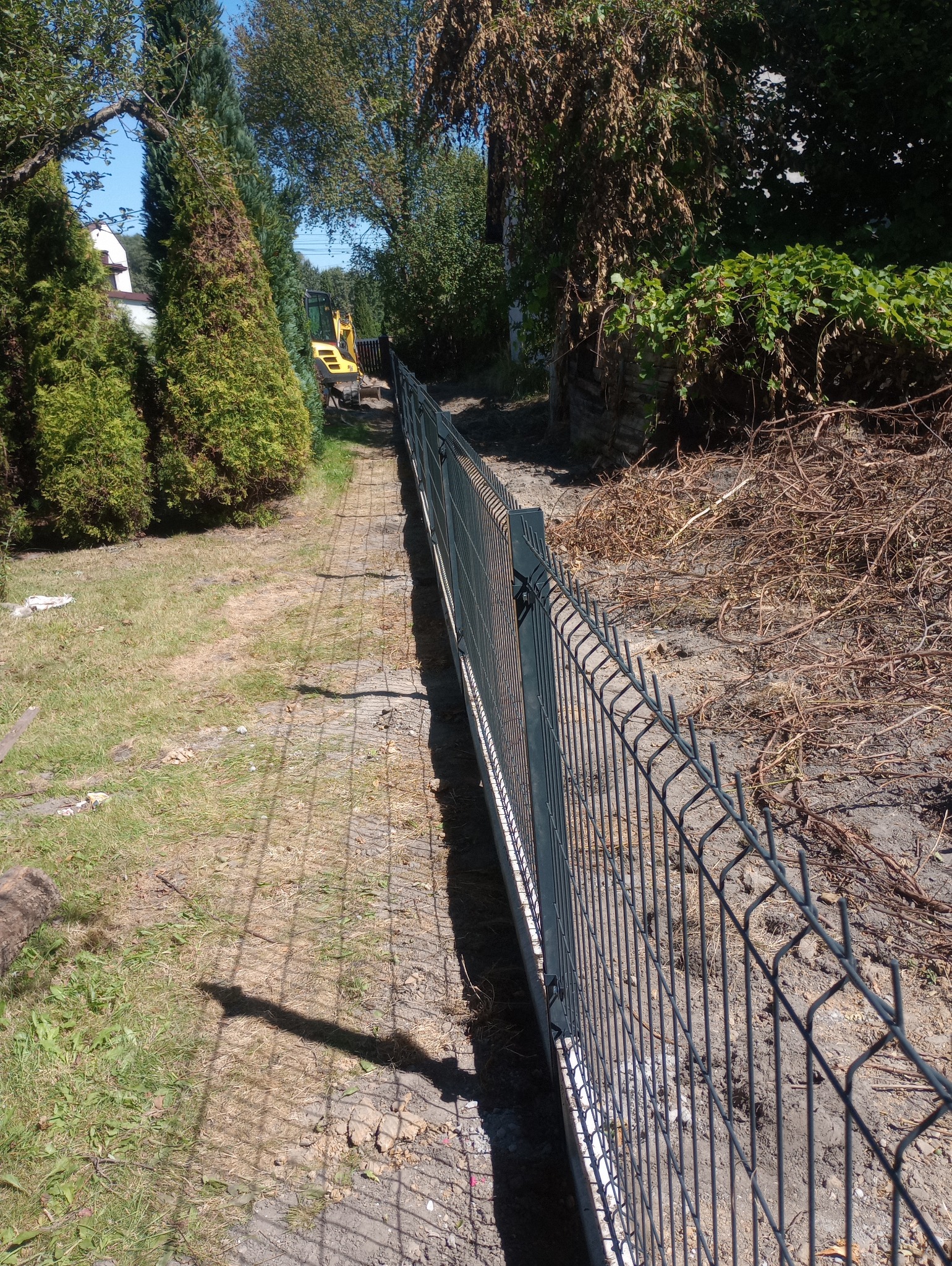 Nowo postawione, metalowe ogrodzenie panelowe wzdłuż wykopu, z widocznym w tle żółtym mini-koparką i zielenią drzew oraz krzewów.