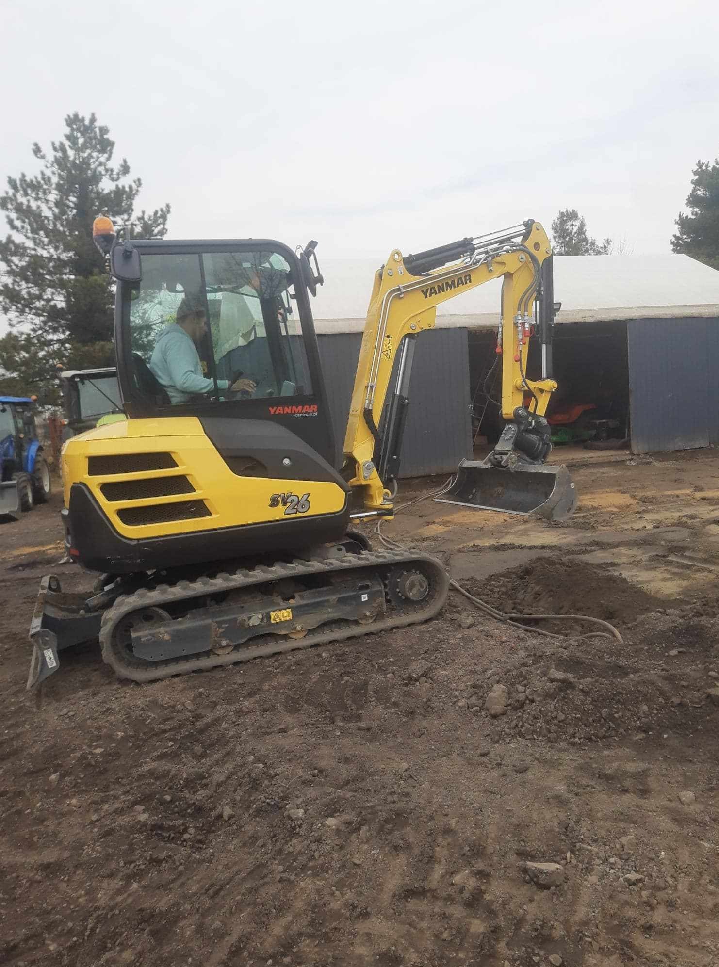 Żółta minikoparka Yanmar SV26 w trakcie pracy, widoczne wykopane zagłębienie w ziemi i wystające rury, operator w kabinie, tło stanowią budynki gospodarcze i drzewa.