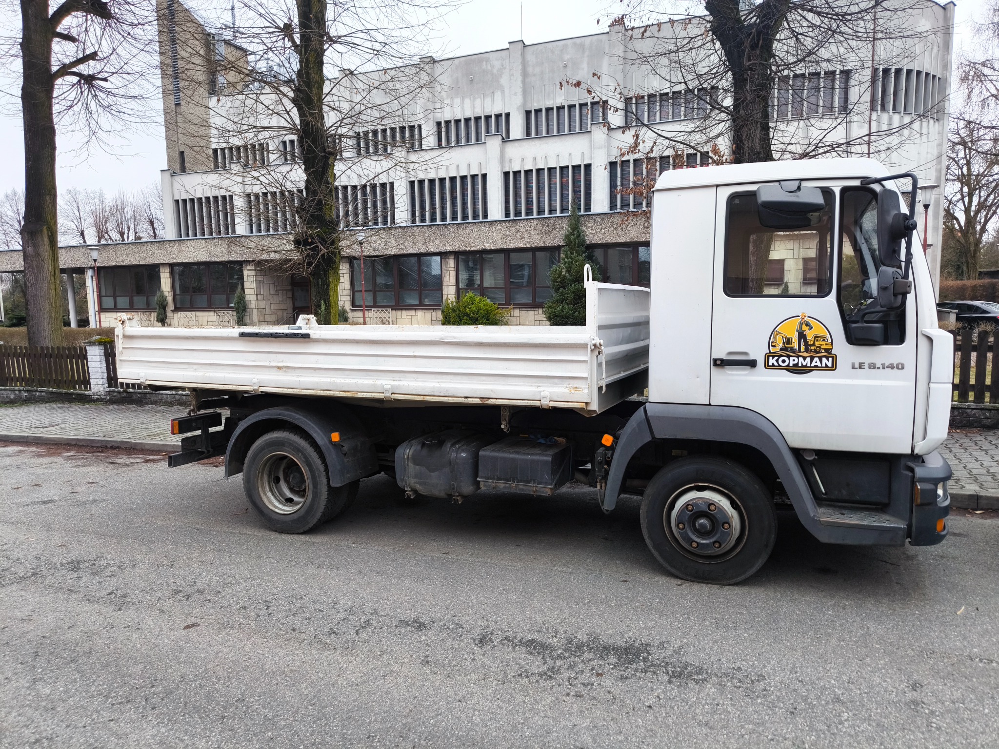 Biała ciężarówka z otwartą skrzynią ładunkową marki Kopman LE 8.140 zaparkowana na ulicy, z widocznym logo firmy na drzwiach kabiny, na tle modernistycznego budynku.