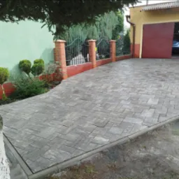 Szara kostka brukowa ułożona w geometryczny wzór, prowadząca do otwartego garażu z widocznym samochodem. Ogród z ogrodzeniem w tle.