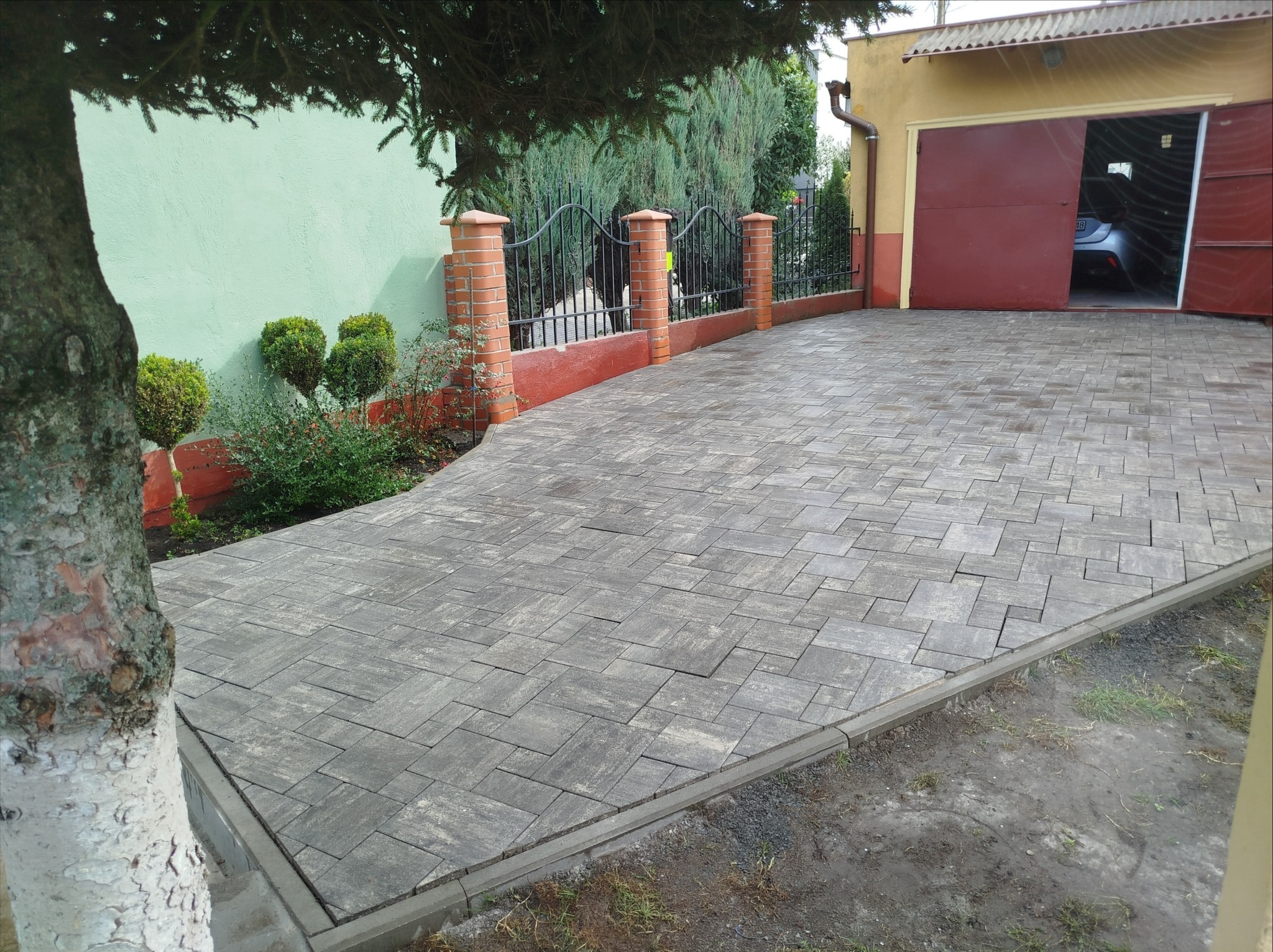 Szara kostka brukowa ułożona w geometryczny wzór, prowadząca do otwartego garażu z widocznym samochodem. Ogród z ogrodzeniem w tle.