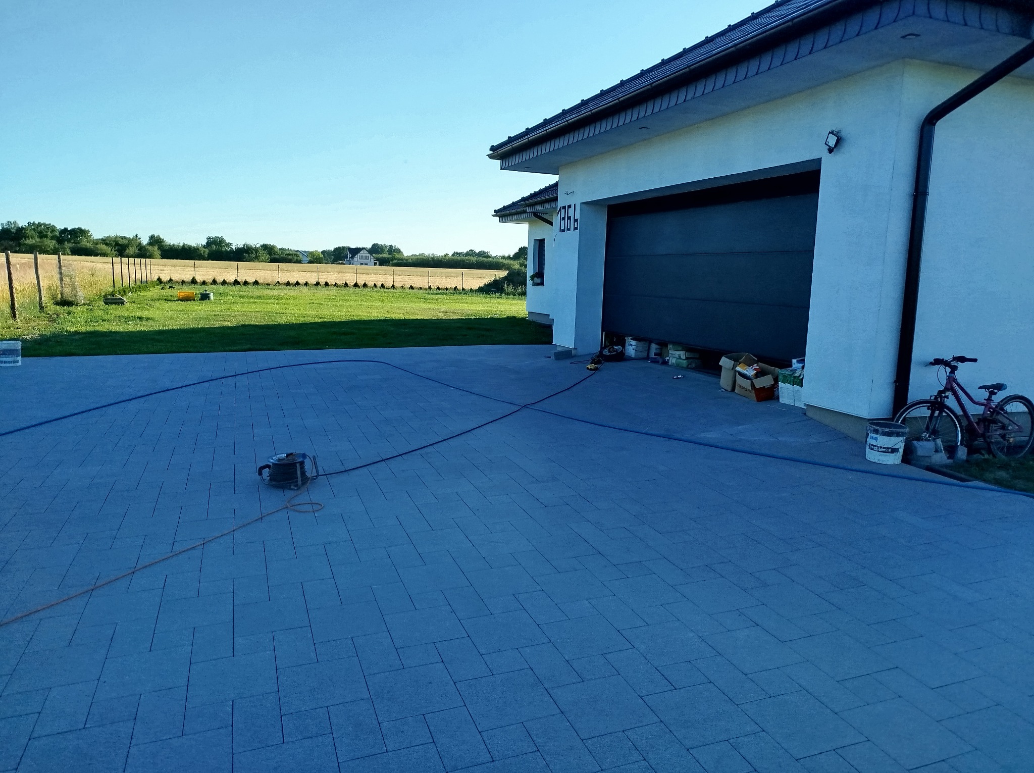 Ułożona, szara kostka brukowa przed domem z otwartym garażem. Widoczny zwinięty kabel i rower oparty o ścianę. W tle trawnik i ogrodzenie.