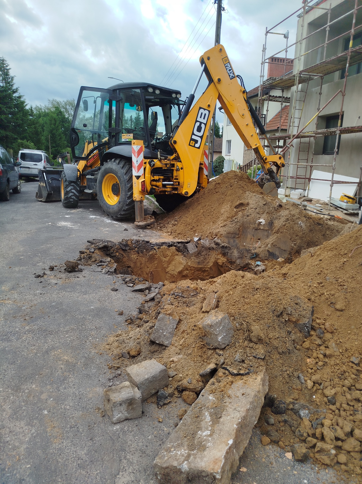 Żółta koparko-ładowarka JCB przy wykopie na asfalcie, obok leżą fragmenty kostki brukowej, w tle budynek w rusztowaniach. Praca w toku.