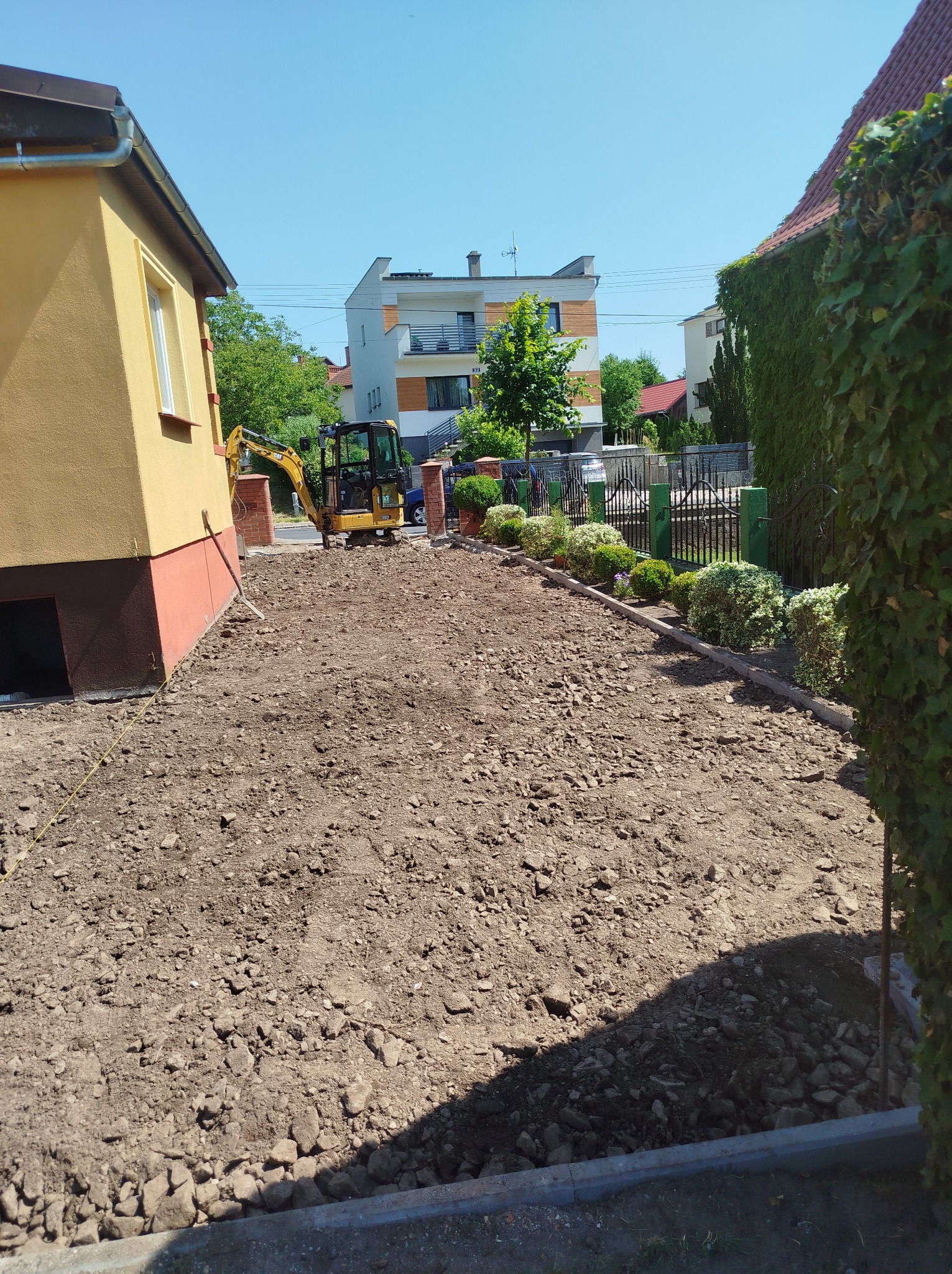 Przygotowany teren pod budowę z widocznym minikoparką CAT i częściowo wykończonym obrzeżem. Słoneczny dzień, widok na domy w tle.