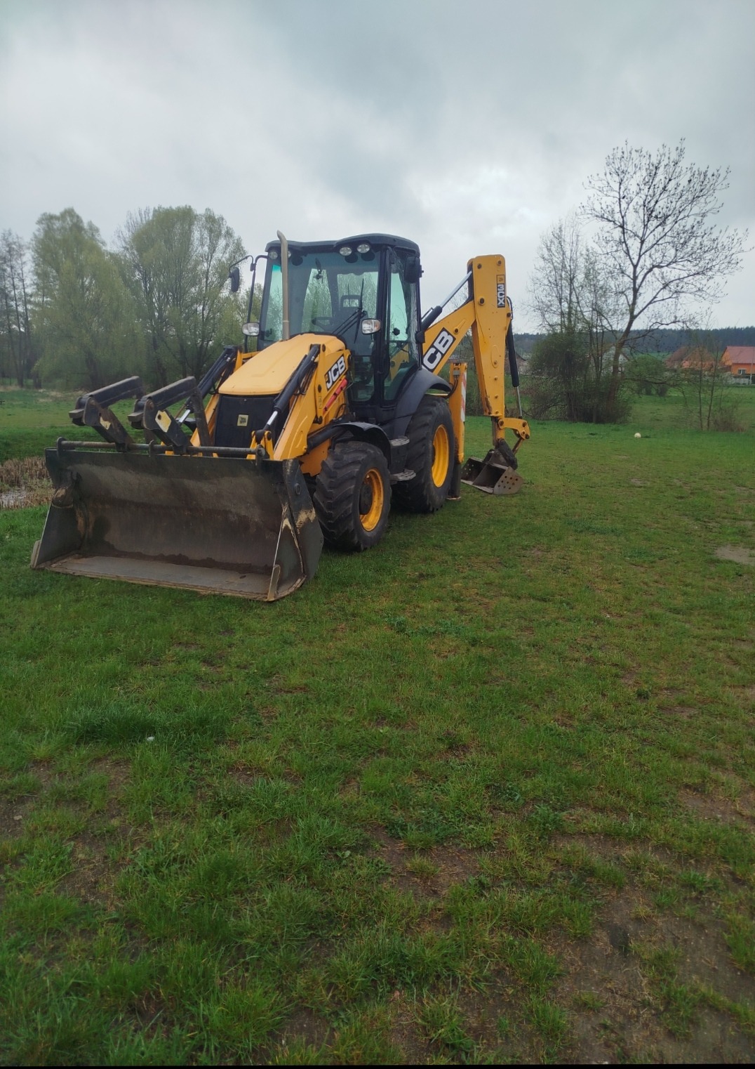 Żółto-czarna koparko-ładowarka JCB na trawiastym terenie, z łyżką skierowaną w stronę widza, gotowa do pracy. W tle drzewa i zabudowania.