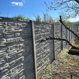 MalMat - Mateusz Pryga - Długi, szary, betonowy płot z fakturą kamienia, widoczny z boku. W tle błękitne niebo i turbina wiatrowa. Na pierwszym planie ziemia i gałęzie drzewa.