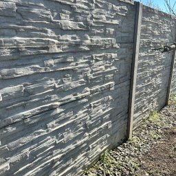 MalMat - Mateusz Pryga - Szary, betonowy płot z teksturą kamienia, widziany pod kątem, z fragmentem nieba i drzew. Płot oddziela teren pokryty kamieniami i ziemią.