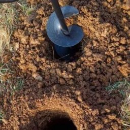 MalMat - Mateusz Pryga - Wiertnica glebowa w akcji, wiercąca otwór w ziemi. Widoczna spirala wiertła i świeżo wykopana ziemia wokół otworu. Przygotowanie pod słupki ogrodzeniowe.