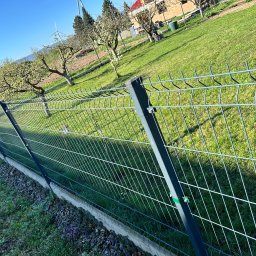 MalMat - Mateusz Pryga - Nowoczesne ogrodzenie panelowe w kolorze antracytowym, oddzielające zielony ogród z drzewami owocowymi i turbinami w tle, wykonane na betonowej podmurówce.