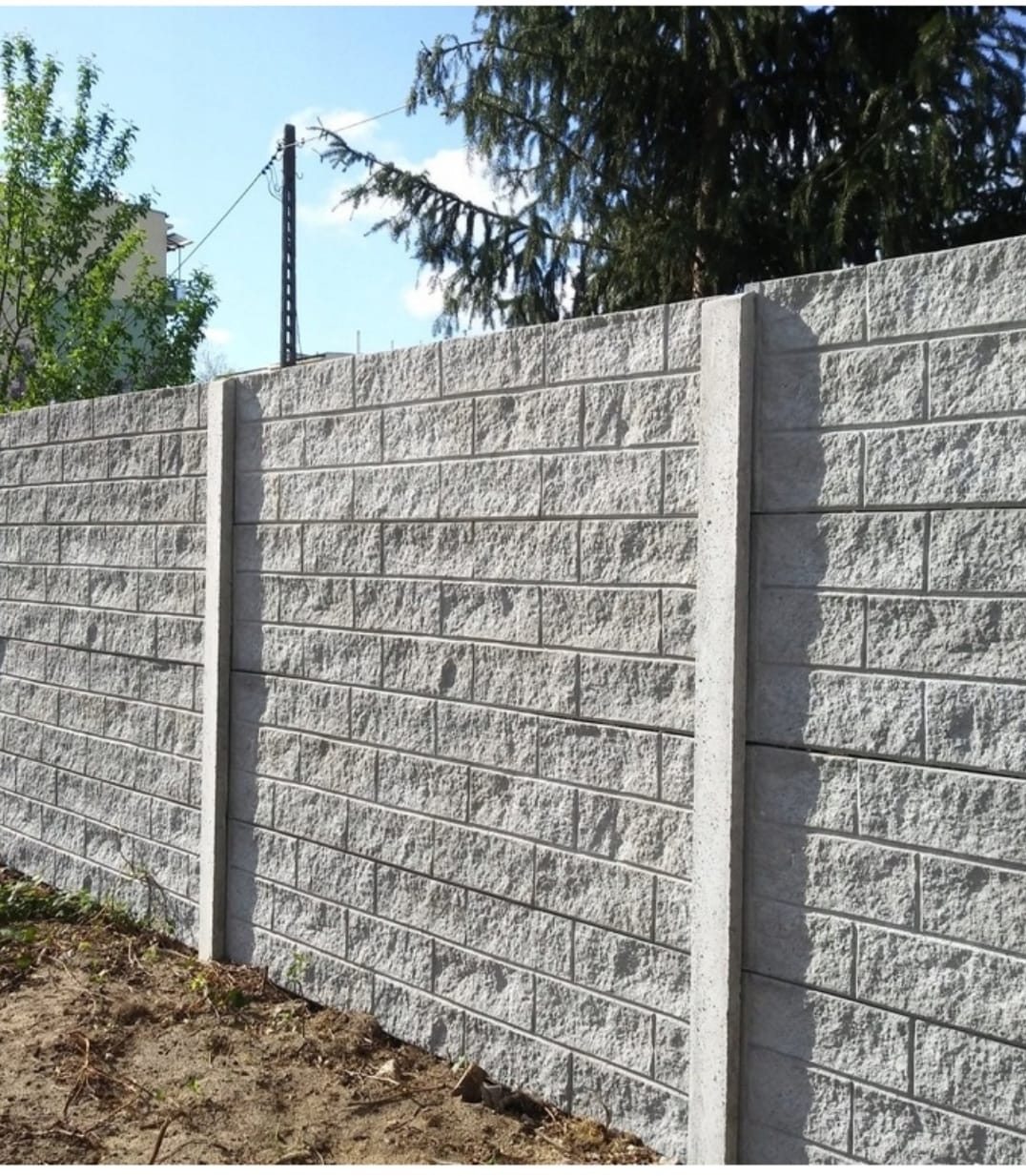 Szary, betonowy mur z teksturą cegieł, wsparty na betonowych słupkach. W tle zieleń drzew i słup energetyczny, błękitne niebo.