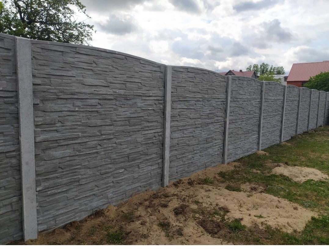 Długi, szary, betonowy płot o fakturze kamienia oddziela posesję. Widoczne słupki i segmenty, tło stanowią domy i pochmurne niebo.