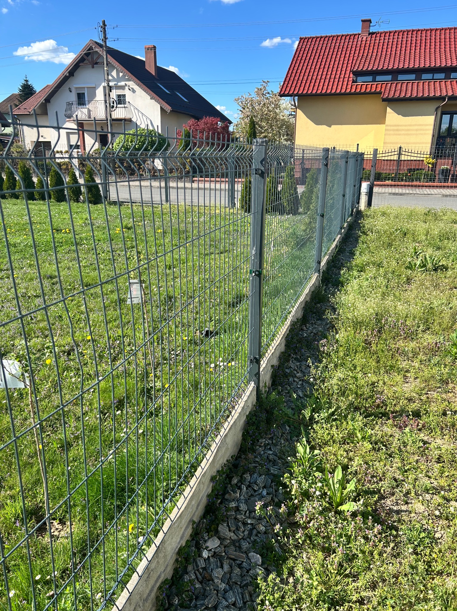 Zielone ogrodzenie panelowe z podmurówką na trawniku, w tle dwa domy jednorodzinne w słoneczny dzień. Widok z poziomu gruntu.