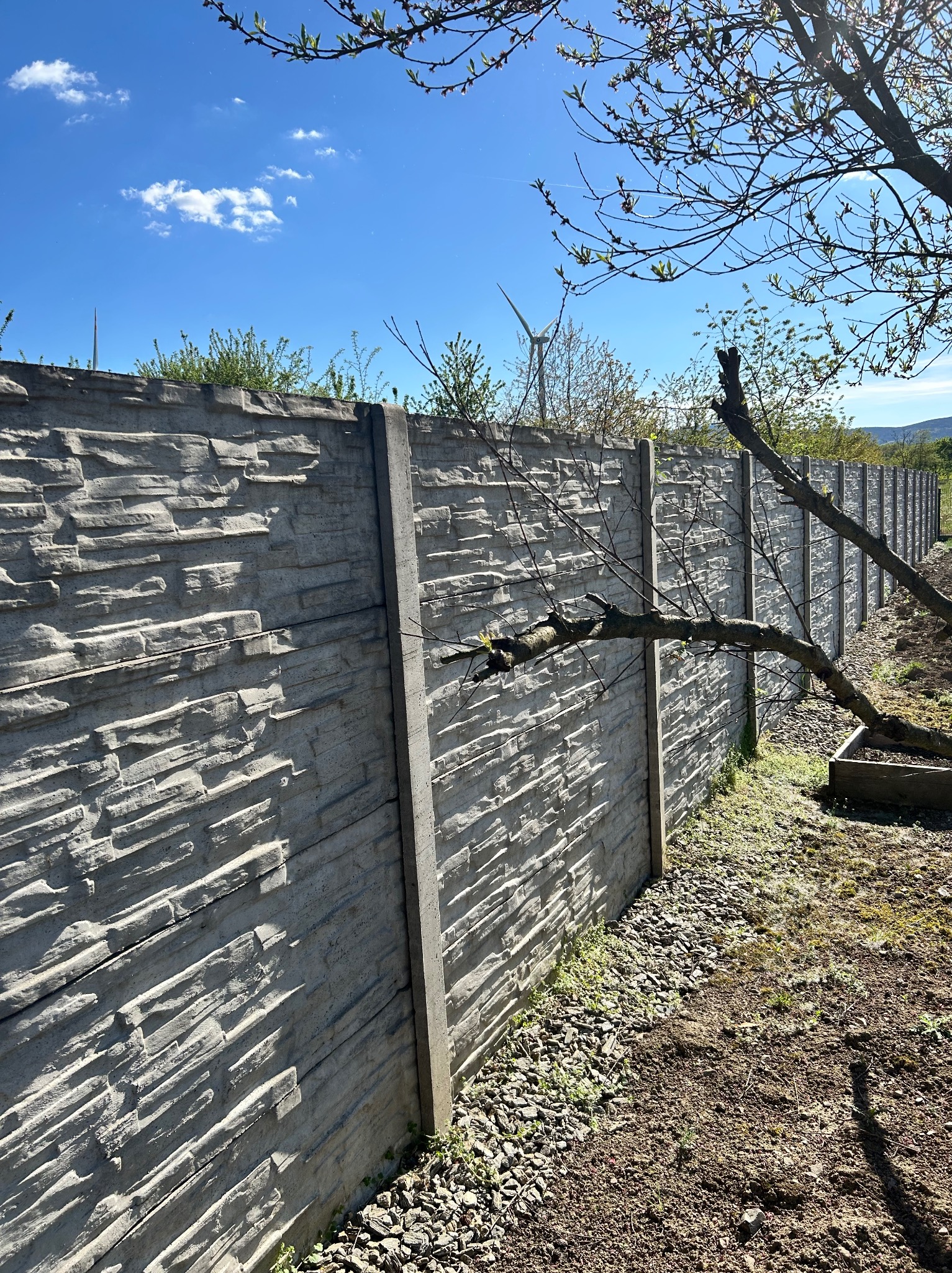 Długi, szary, betonowy płot z fakturą kamienia, widoczny z boku. W tle błękitne niebo i turbina wiatrowa. Na pierwszym planie ziemia i gałęzie drzewa.