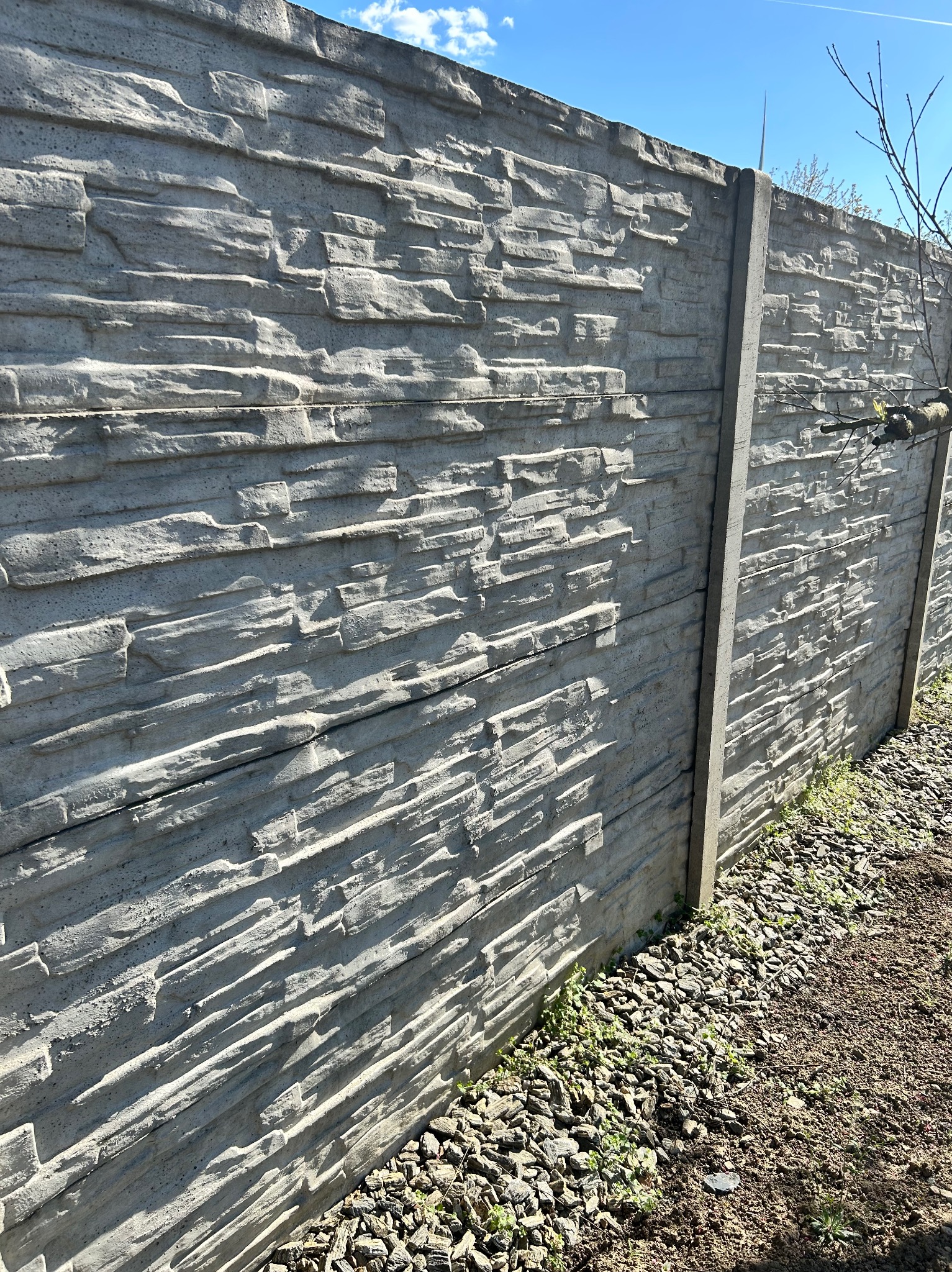 Szary, betonowy płot z teksturą kamienia, widziany pod kątem, z fragmentem nieba i drzew. Płot oddziela teren pokryty kamieniami i ziemią.
