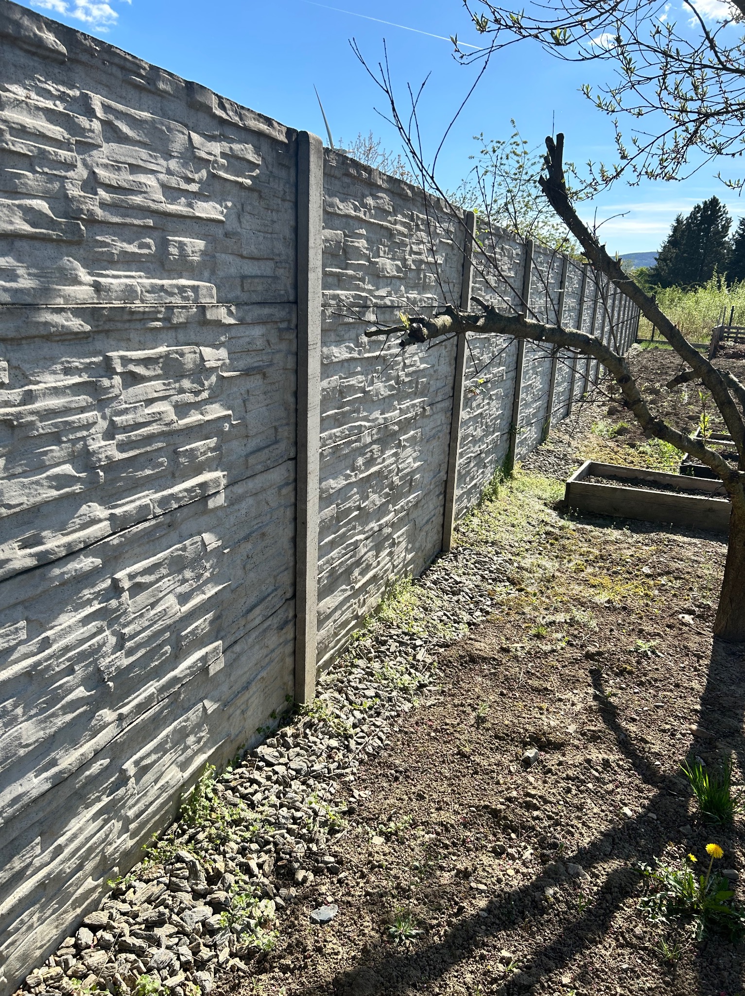 Długi, szary mur z betonowych płyt imitujących kamień, oddzielający ogród. Widoczne słupki, kamienny pas u podstawy i fragment drzewa z prawej strony.
