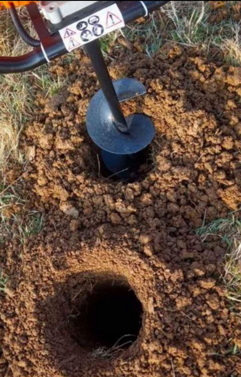 Wiertnica glebowa w akcji, wiercąca otwór w ziemi. Widoczna spirala wiertła i świeżo wykopana ziemia wokół otworu. Przygotowanie pod słupki ogrodzeniowe.