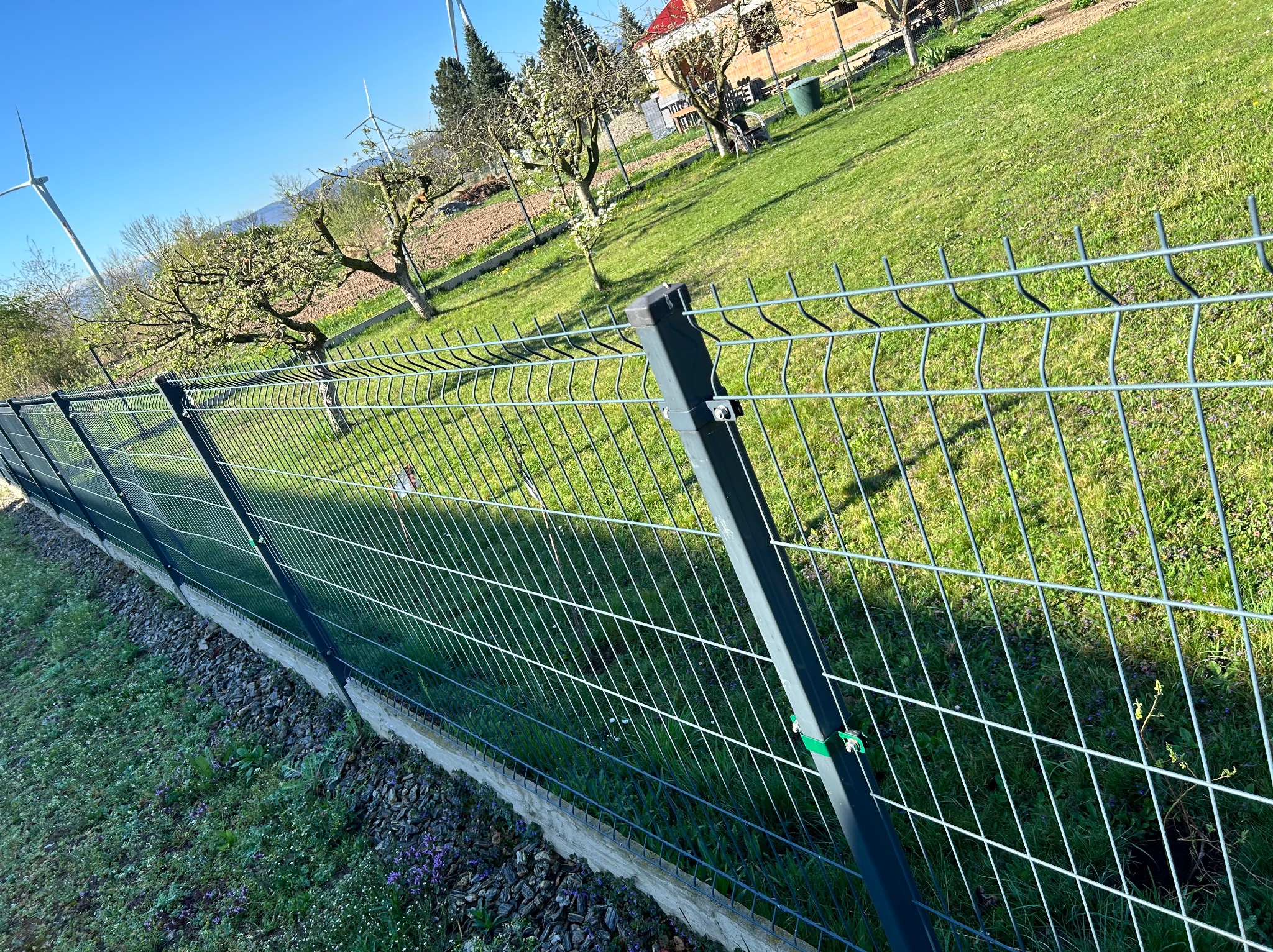 Nowoczesne ogrodzenie panelowe w kolorze antracytowym, oddzielające zielony ogród z drzewami owocowymi i turbinami w tle, wykonane na betonowej podmurówce.