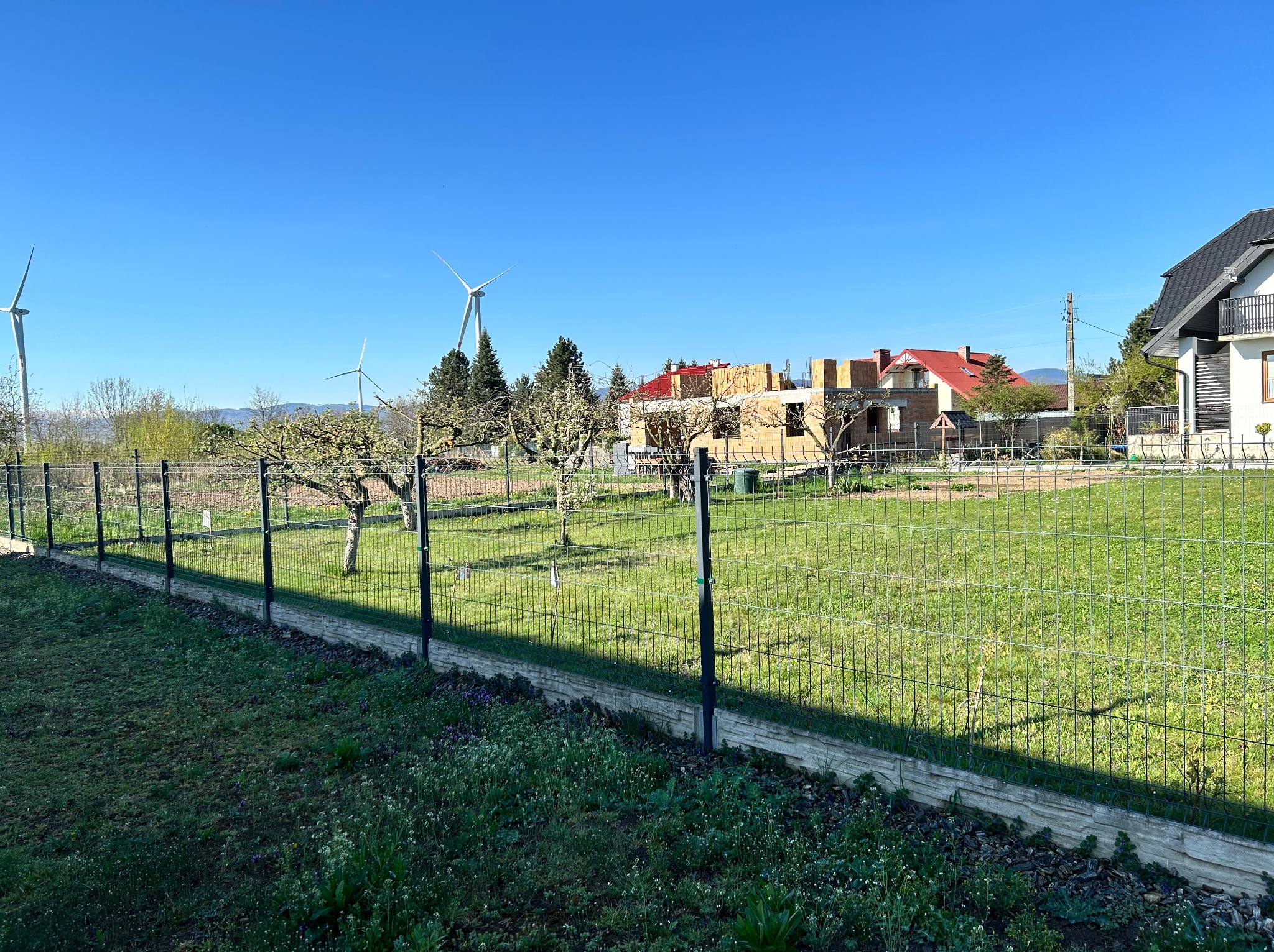 Nowoczesne ogrodzenie panelowe na tle zielonego trawnika, w oddali budowa domu i turbiny wiatrowe pod błękitnym niebem. Słoneczny dzień.