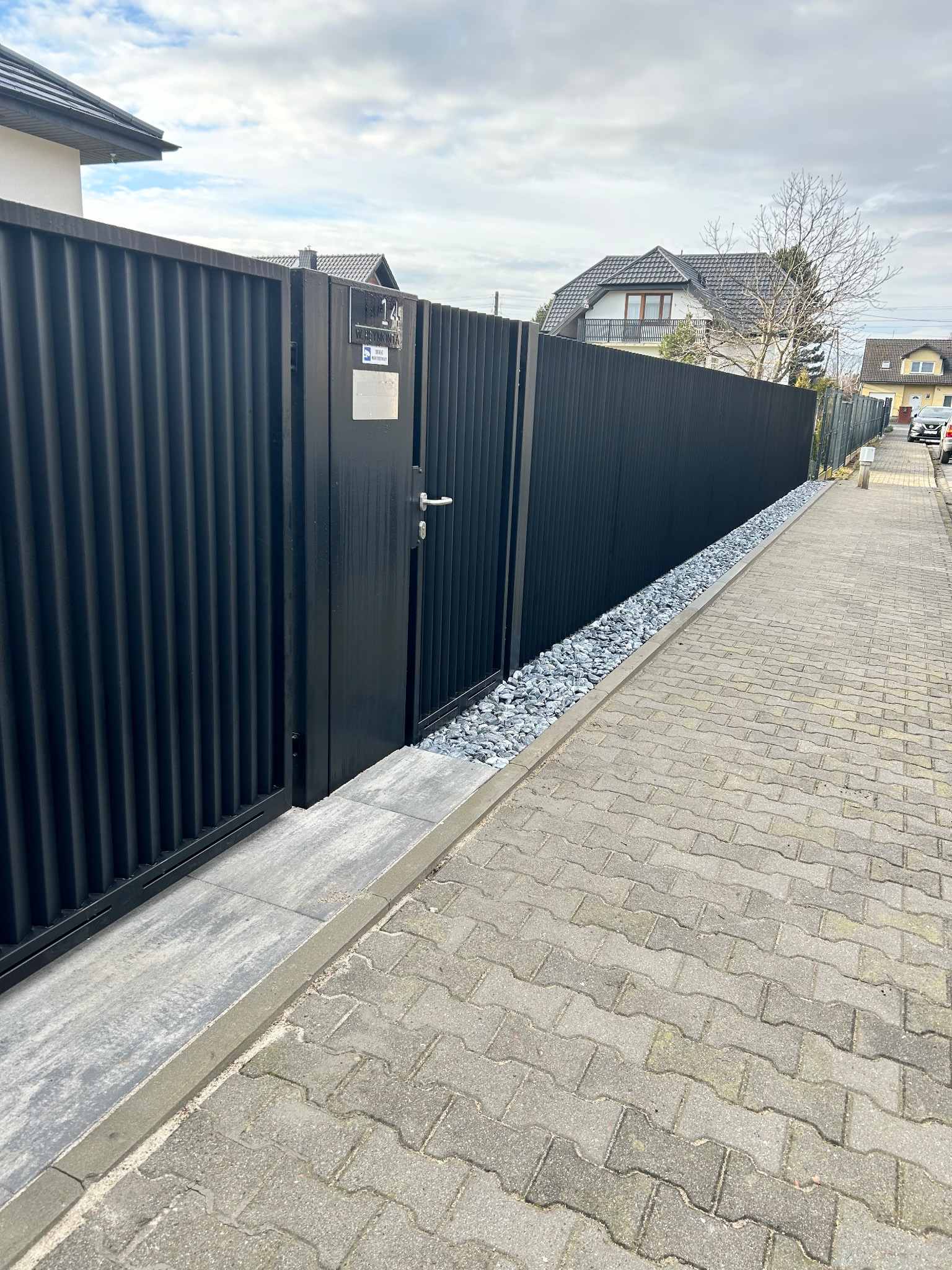 Nowoczesne, czarne ogrodzenie panelowe z bramką, wykończone szarym kamieniem. Widok z boku, na tle kostki brukowej i zabudowań.