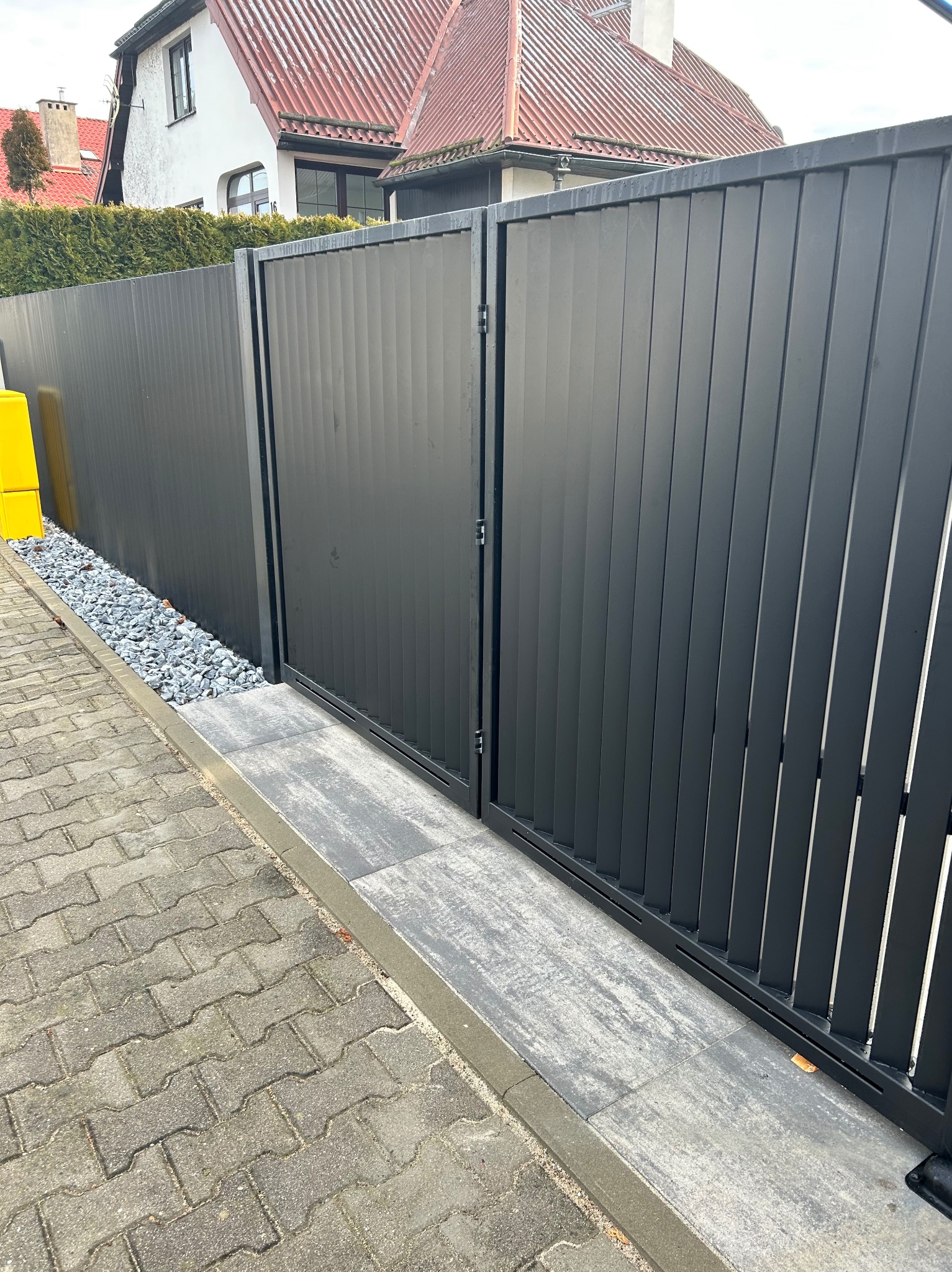 Nowoczesna, grafitowa brama panelowa z pionowymi szczeblami, zamontowana na posesji z kostką brukową i kamiennym podsypaniem. Minimalistyczny design.